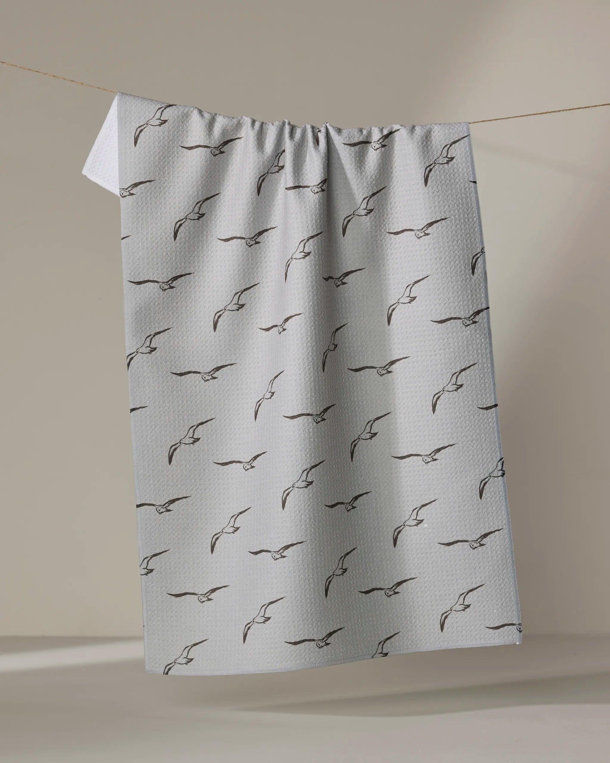 The Jenny Keller Shoreline Scout is a light gray absorbent dish towel featuring a pattern of simple black outlined birds in flight, shown hanging neatly from a clothesline against a neutral background.