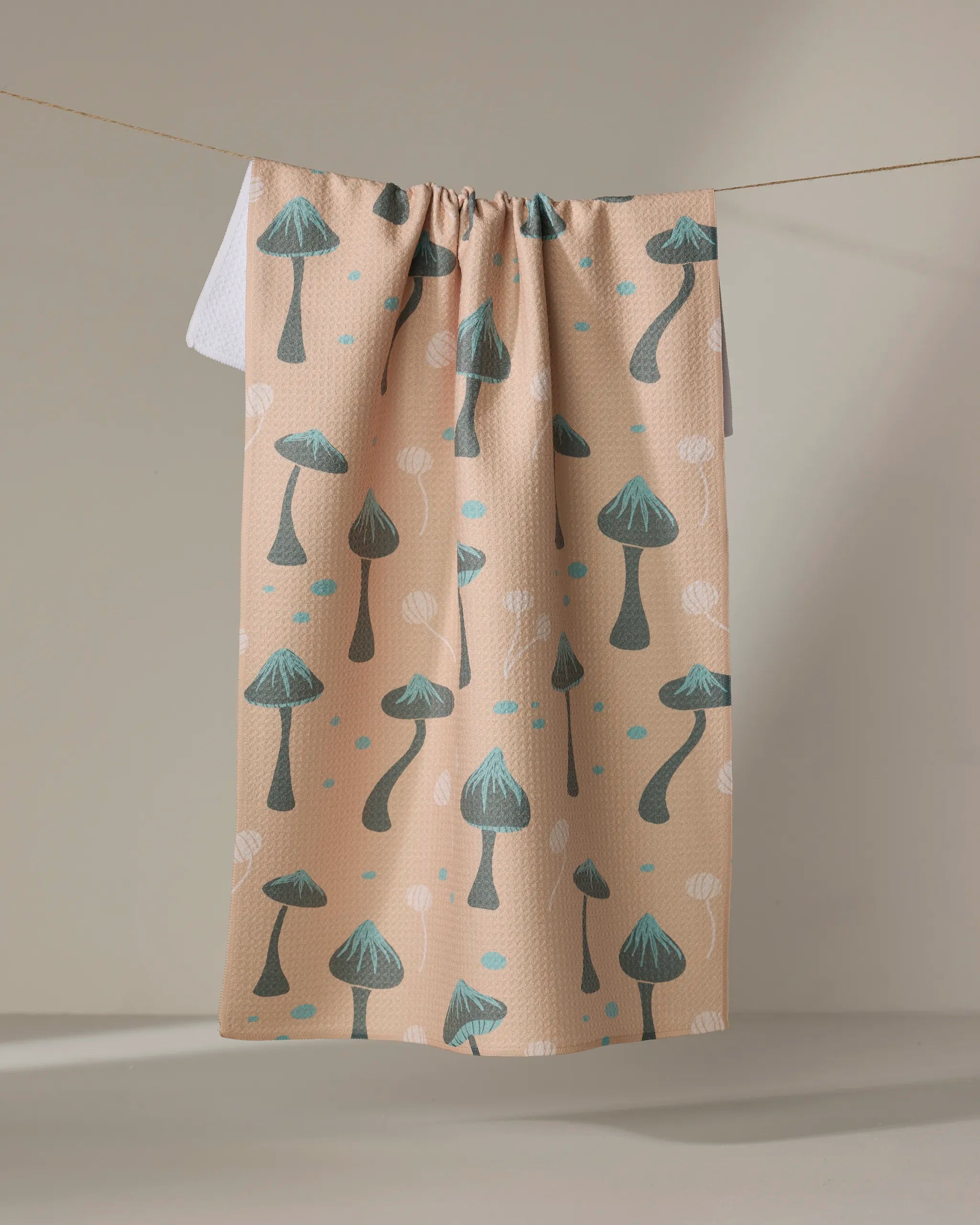 The Amberleigh Adoff Shroom Sprouts dish towel, featuring teal and dark gray mushrooms with blue dots on beige fabric, hangs on a line with a plain white tea towel partially visible behind it.