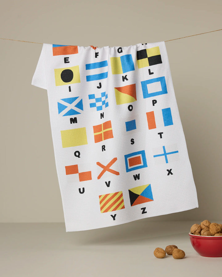 The Geometry Signal Flags dish towel, featuring the alphabet and vibrant nautical signal flags, hangs on a string, with a small red bowl of walnuts resting nearby on a beige surface.
