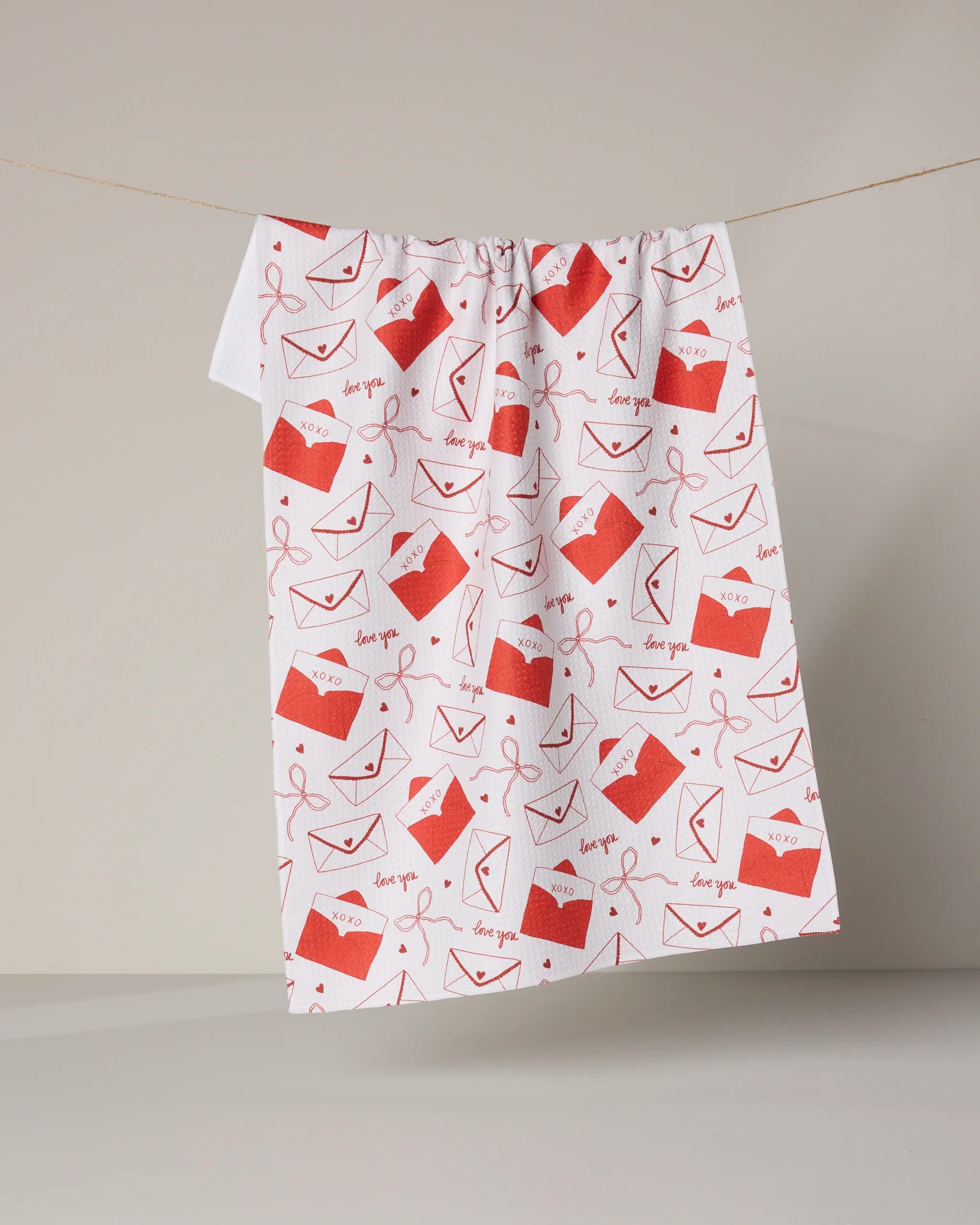 The Geometry "Signed" tea towel, ultra-absorbent and white with red envelope, heart, and "love you" designs, is displayed hanging against a plain beige background.