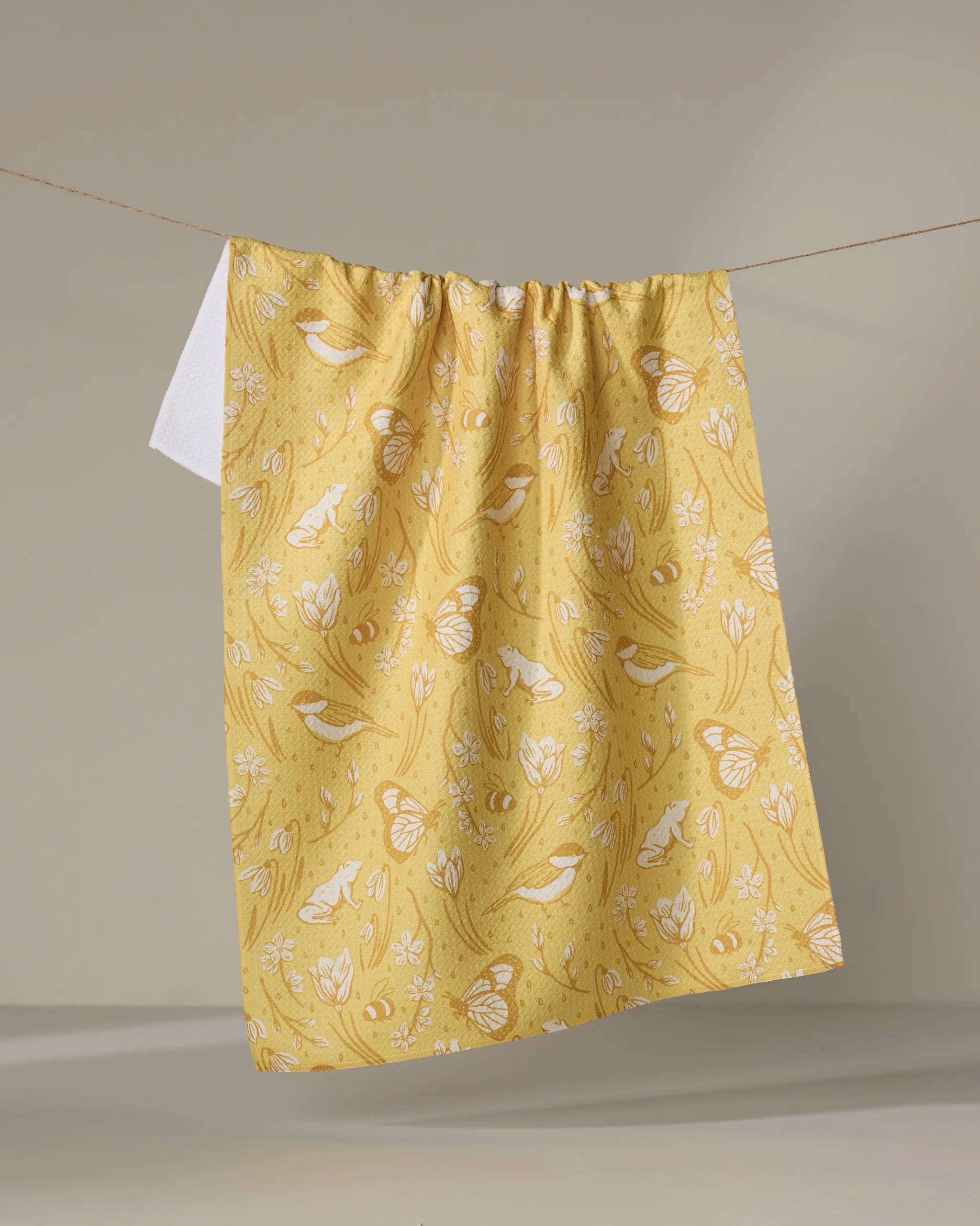 The "Signs of Spring" tea towel by Sarah Daugherty features a white nature-inspired bird, butterfly, and leaf pattern on yellow and hangs on a clothesline against a neutral background.