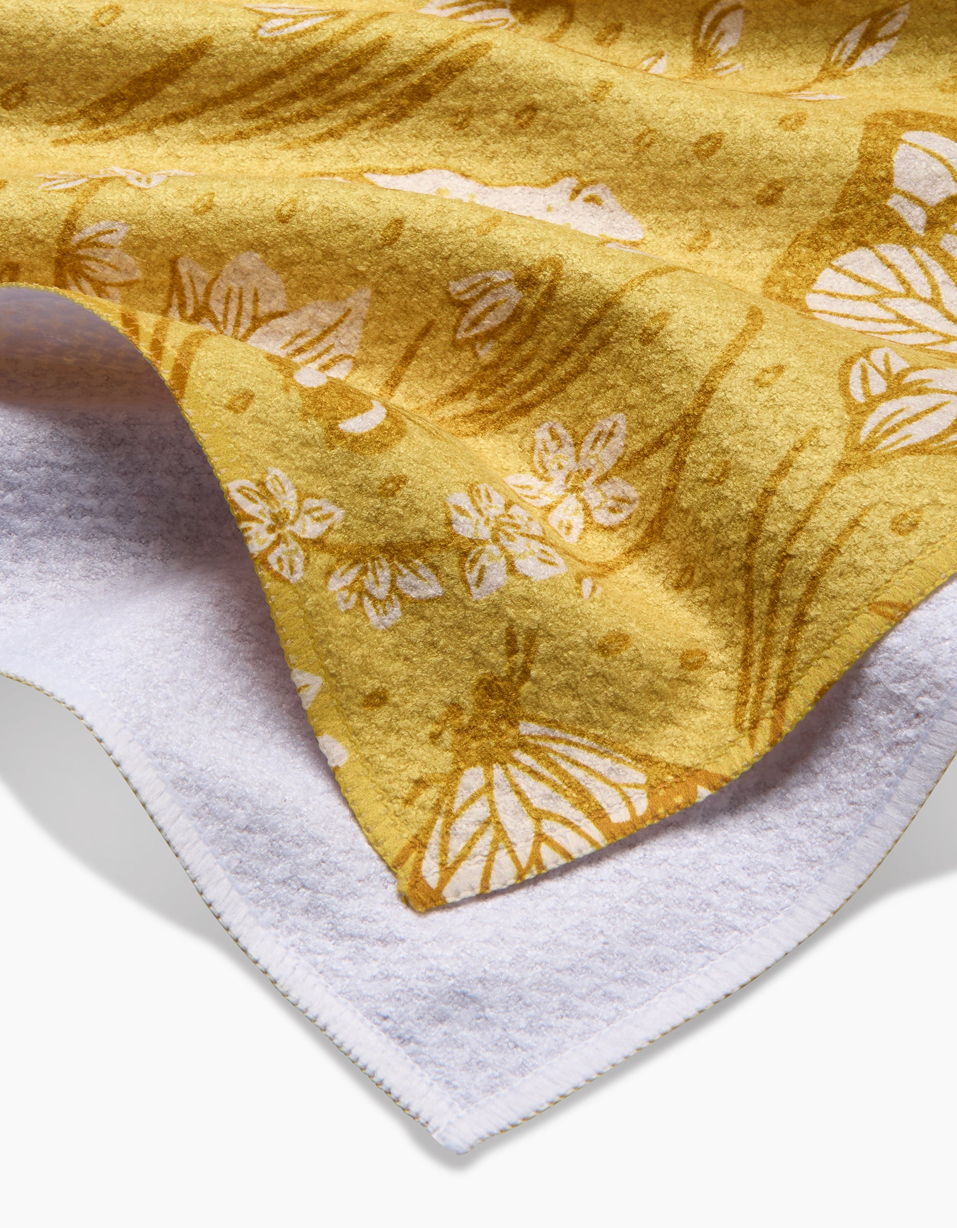 A close-up of the Signs of Spring tea towel by Sarah Daugherty, made from ultra-absorbent Geoweave fabric, featuring soft yellow tones with white and brown floral and animal patterns, draped over a white textured surface.