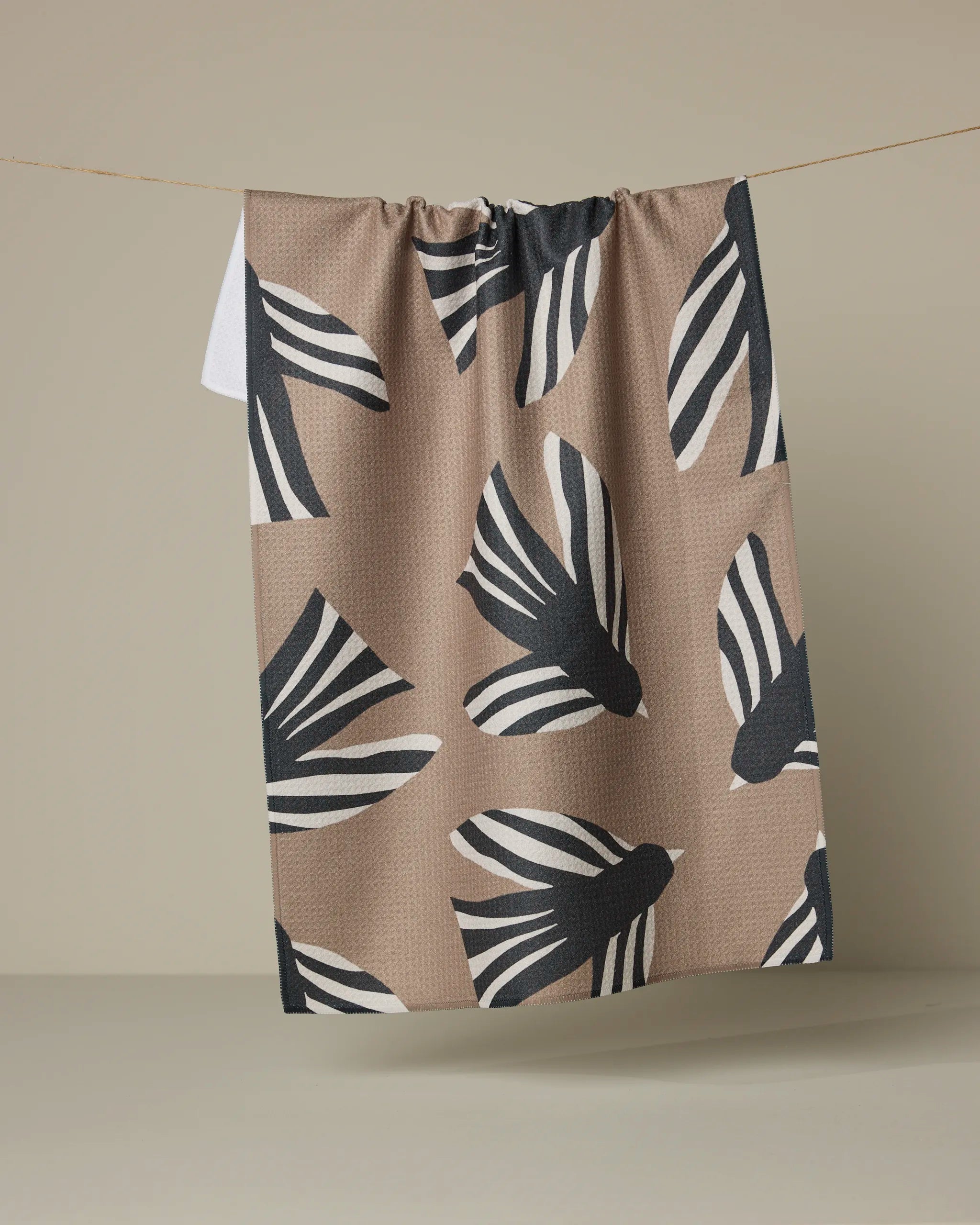 The Anna Christian Sigrid tea towel, crafted from recycled materials in tan and adorned with bold black and white bird motifs, hangs on a thin string against a neutral beige background.