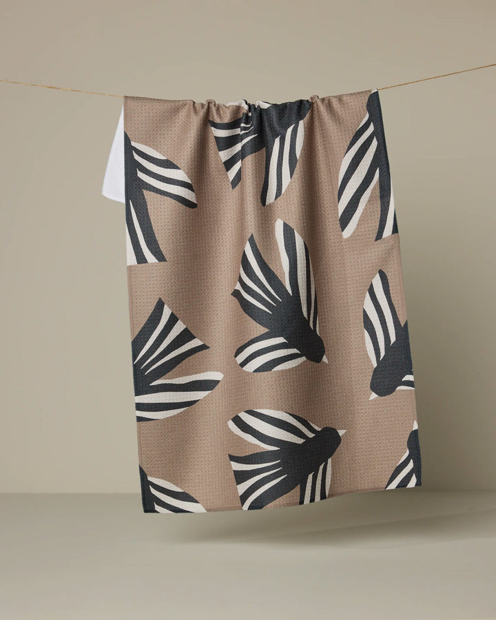 The Anna Christian Sigrid tea towel, crafted from recycled materials in tan and adorned with bold black and white bird motifs, hangs on a thin string against a neutral beige background.