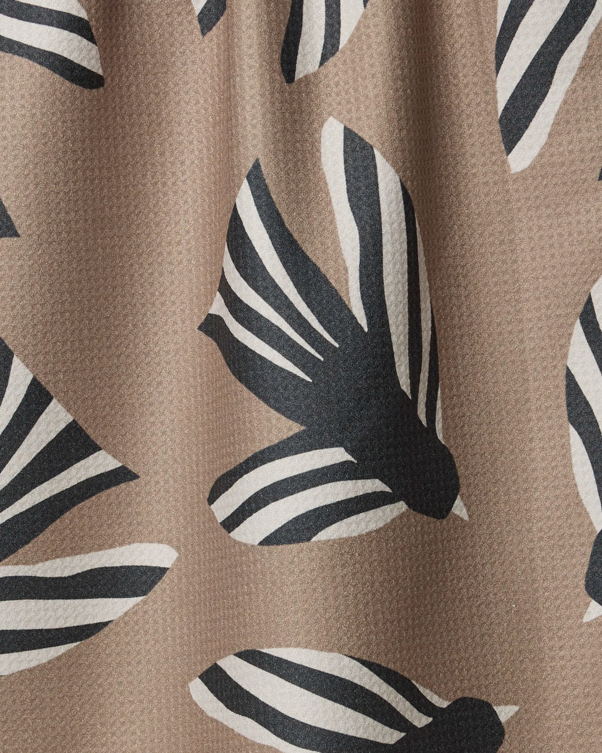 A close-up of the Sigrid tea towel by Anna Christian, made from recycled ultra-absorbent brown fabric, showcases a repeating pattern of stylized black and white birds with striped wings and visible texture.