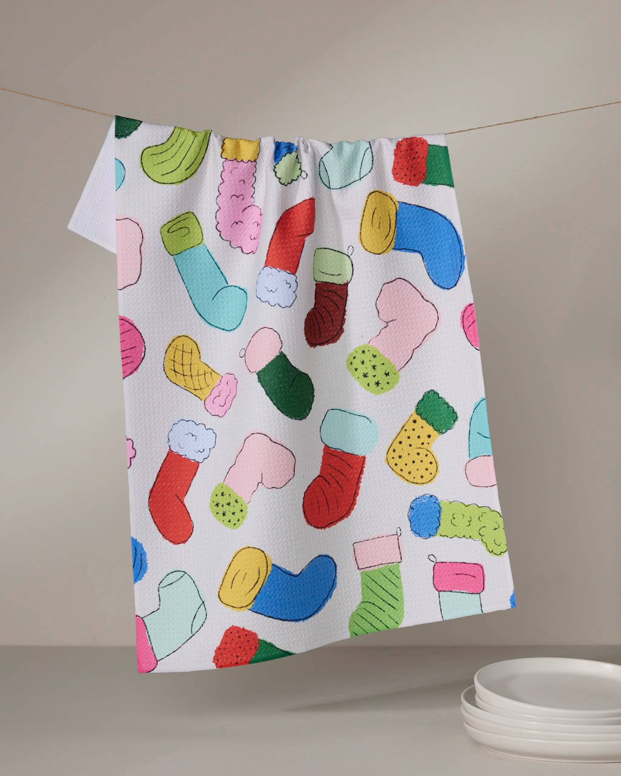 The Geometry Silly Stockings tea towel features a colorful Christmas stocking pattern on ultra-absorbent fabric, adding festive cheer to your kitchen as it hangs above a stack of white plates against a neutral background.