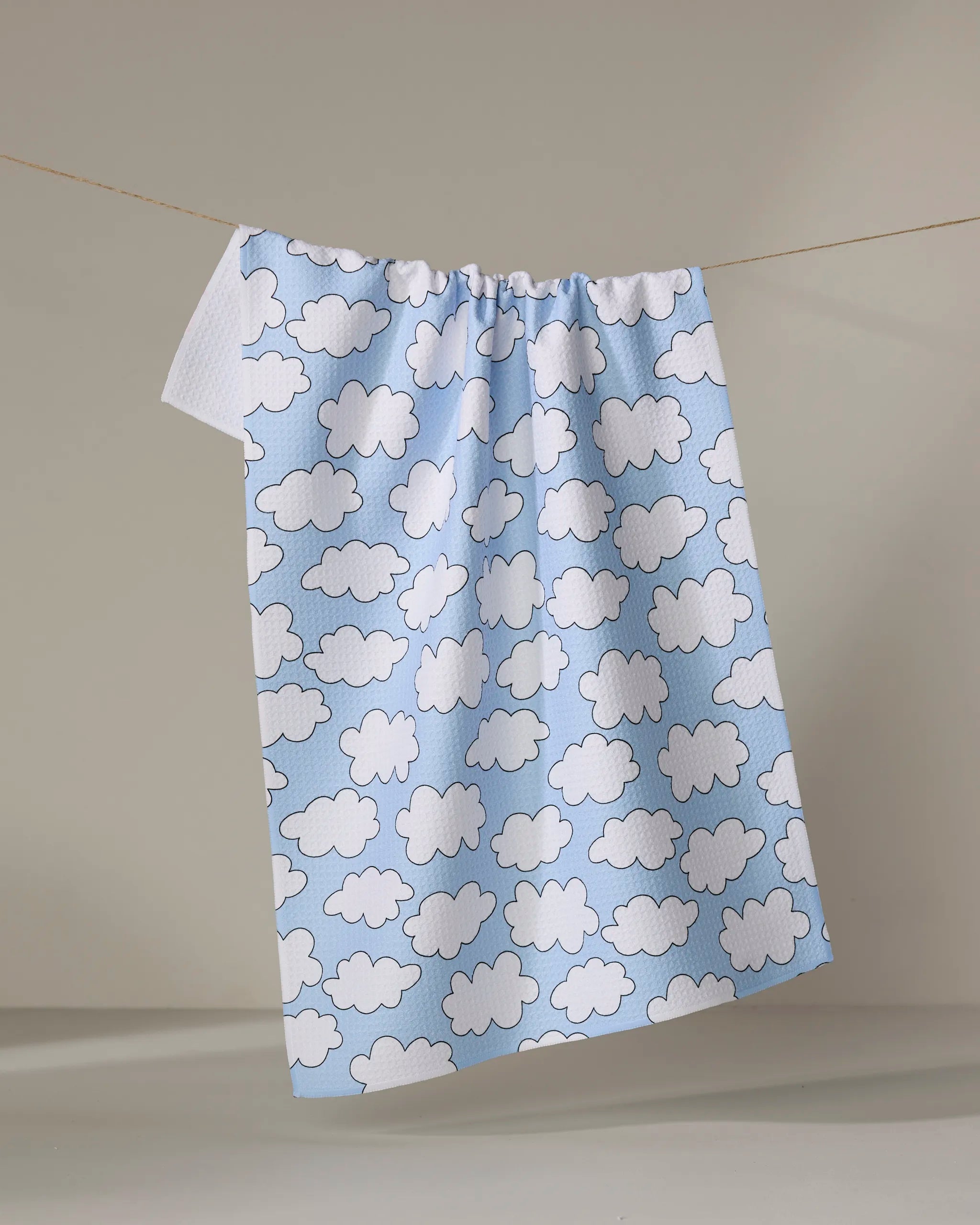 The Silver Linings towel by Mindfulnice, made from recycled materials, features a light blue color with a white cloud pattern and ultra-absorbent design, shown hanging on a clothesline against a neutral background.