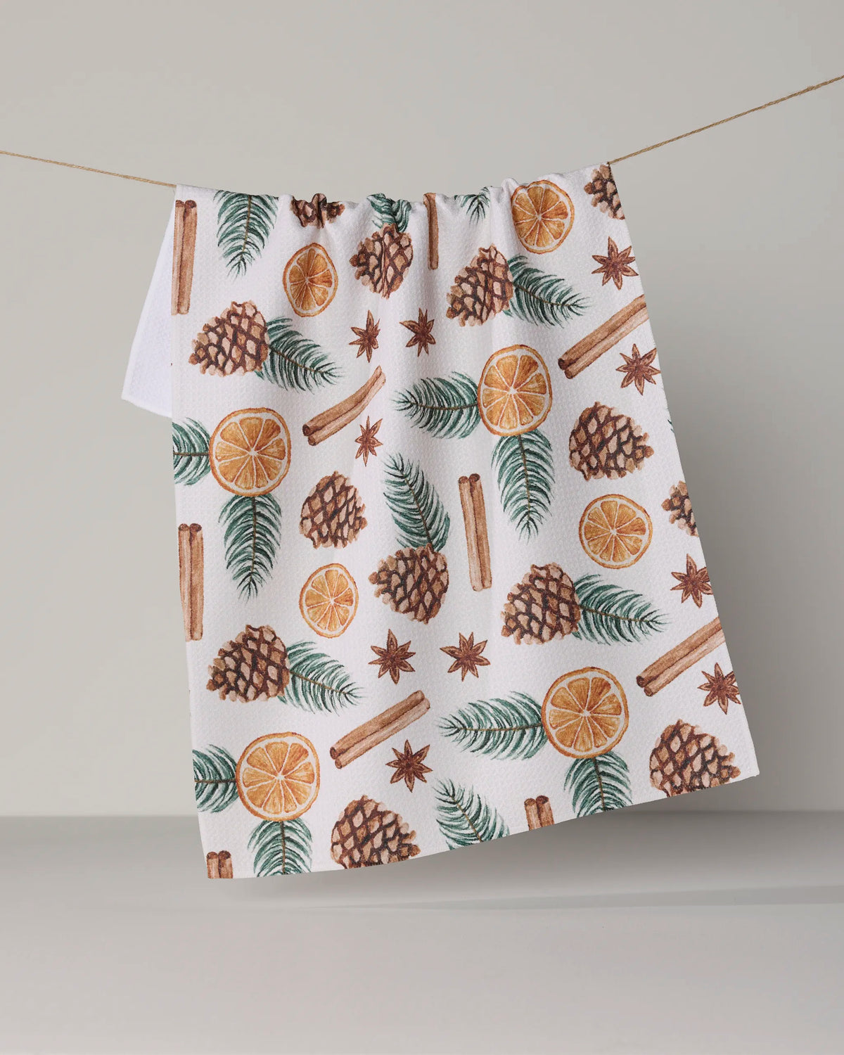 A Geometry Simmer Pot kitchen towel hangs on a line, featuring a charming pattern of pinecones, orange slices, cinnamon sticks, star anise, and green leaves against a crisp white background.