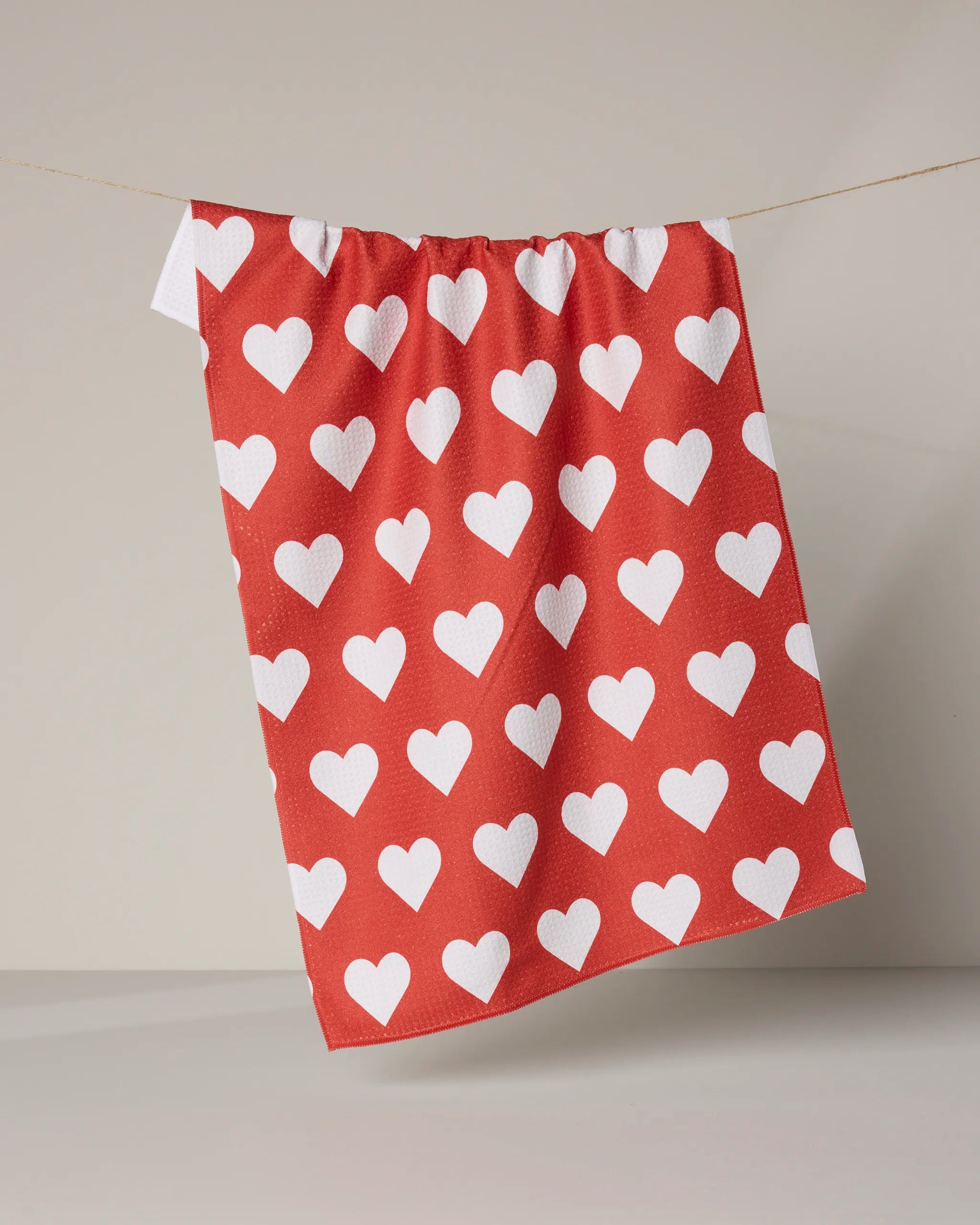 The Geometry Simply Love dish towel, red with evenly spaced white hearts, hangs on a clothesline against a plain, light background.