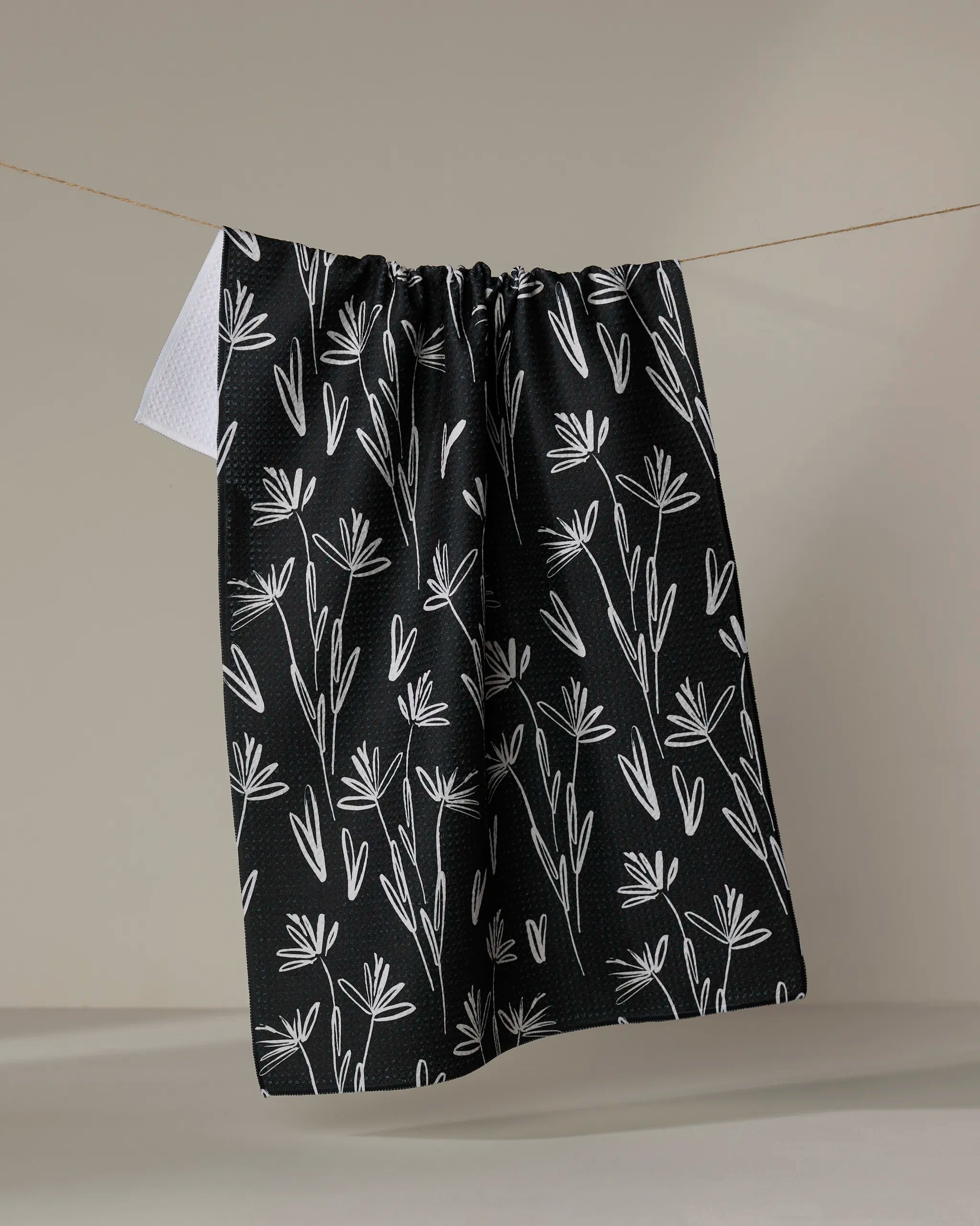 The Toboggan Avenue Sketchbook Dust tea towel, featuring a white floral pattern on black, hangs on a clothesline against a neutral background with one white corner of the cloth partially tucked behind.