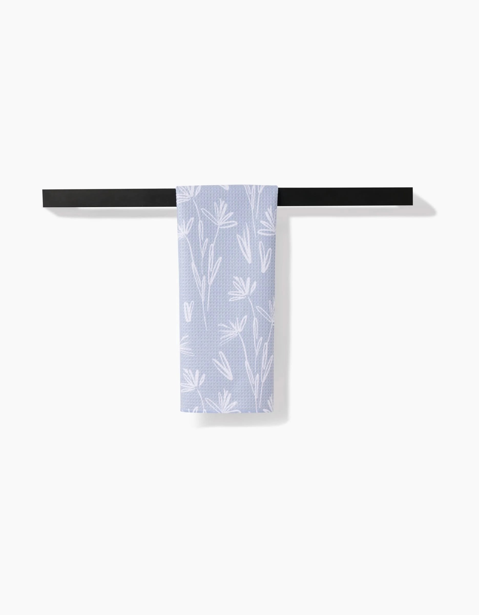 A light purple, ultra-absorbent Toboggan Avenue Sketchbook Floral towel with a white floral pattern hangs neatly over a modern black horizontal towel rack against a plain white background.