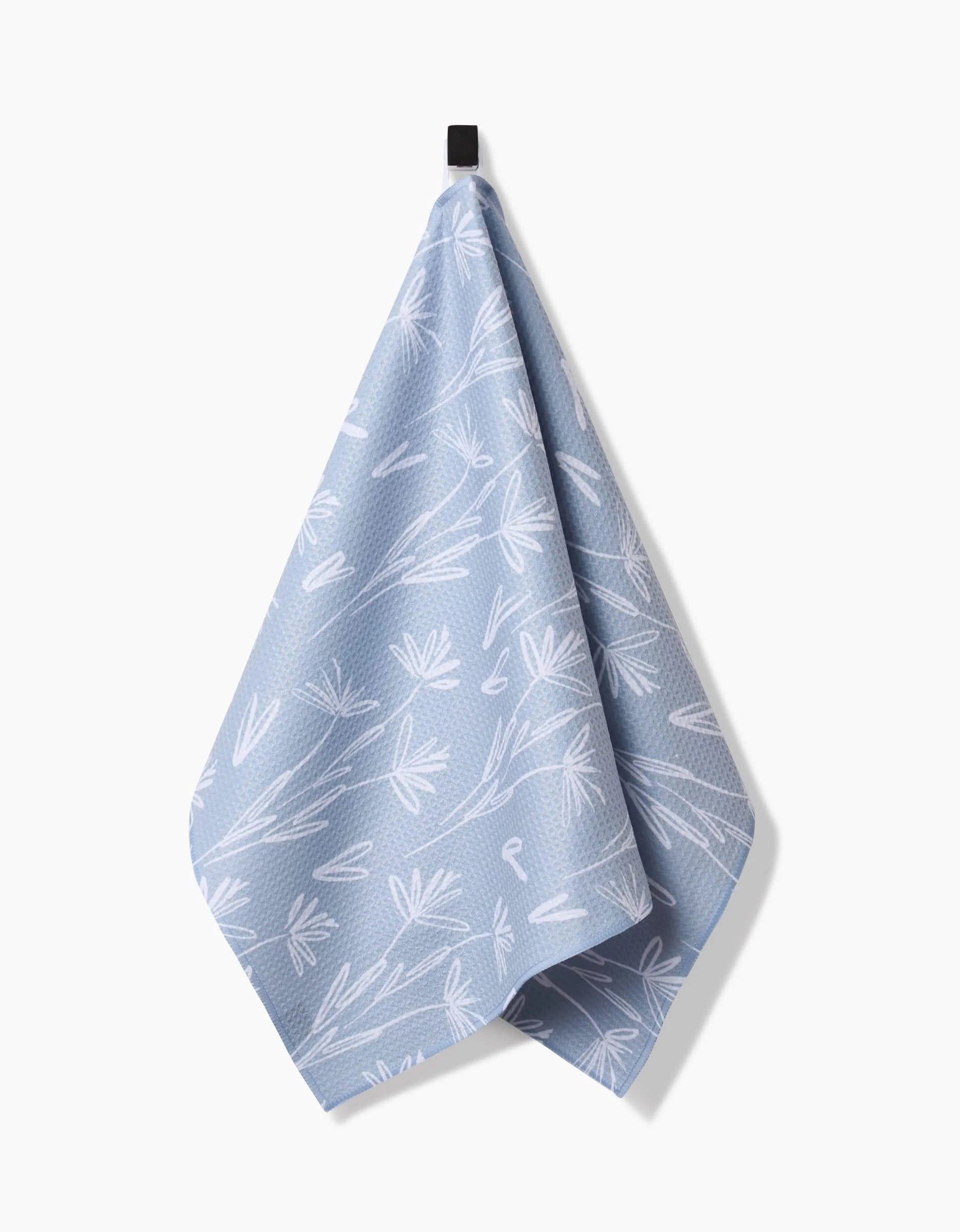 A light blue ultra-absorbent Sketchbook Floral tea towel by Toboggan Avenue, featuring a white floral pattern, hangs from a black loop against a plain white background.