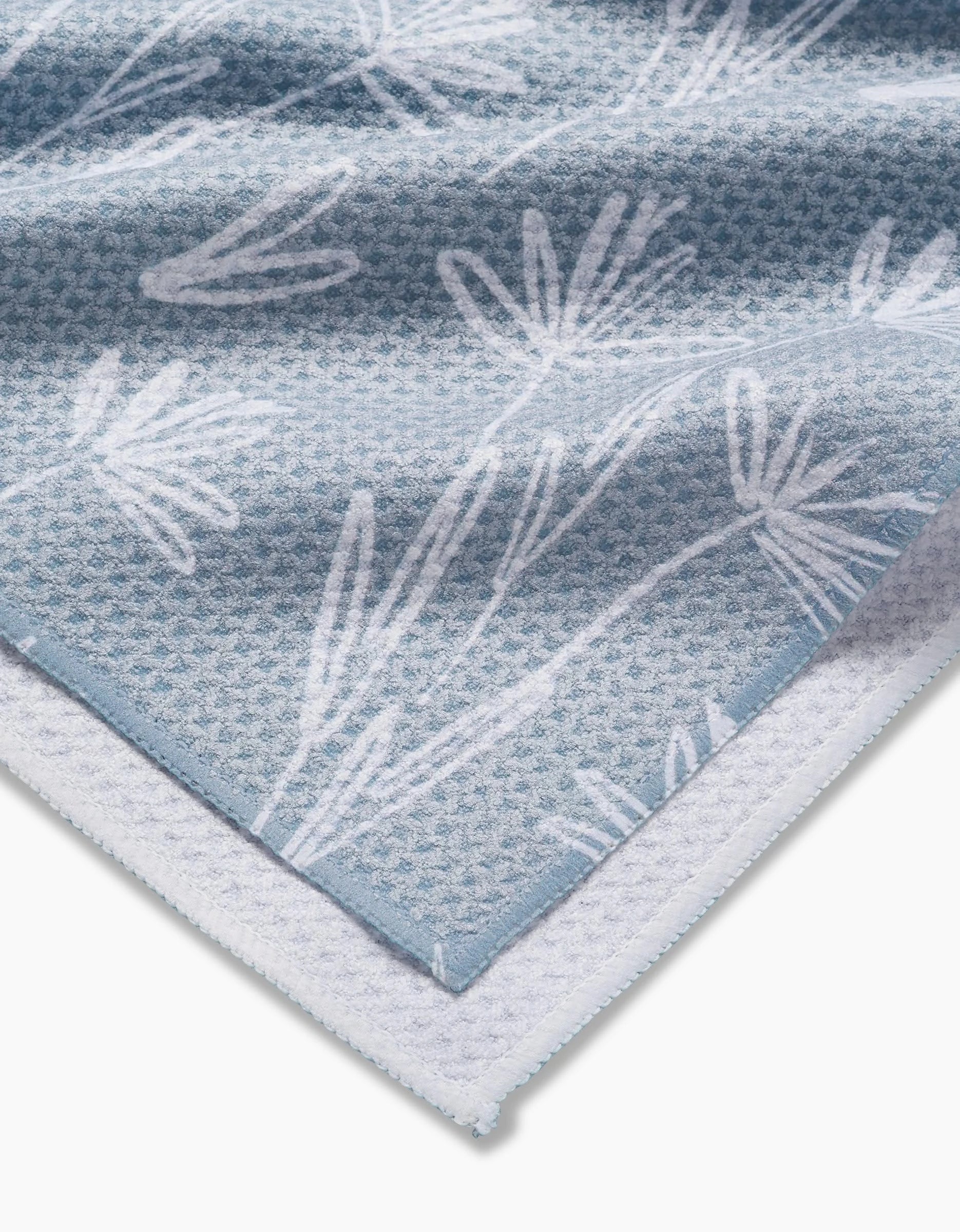 A close-up of the Sketchbook Floral tea towel by Toboggan Avenue, showing its blue and white recycled material with a subtle waffle pattern, floral design, and a folded corner revealing the white underside.