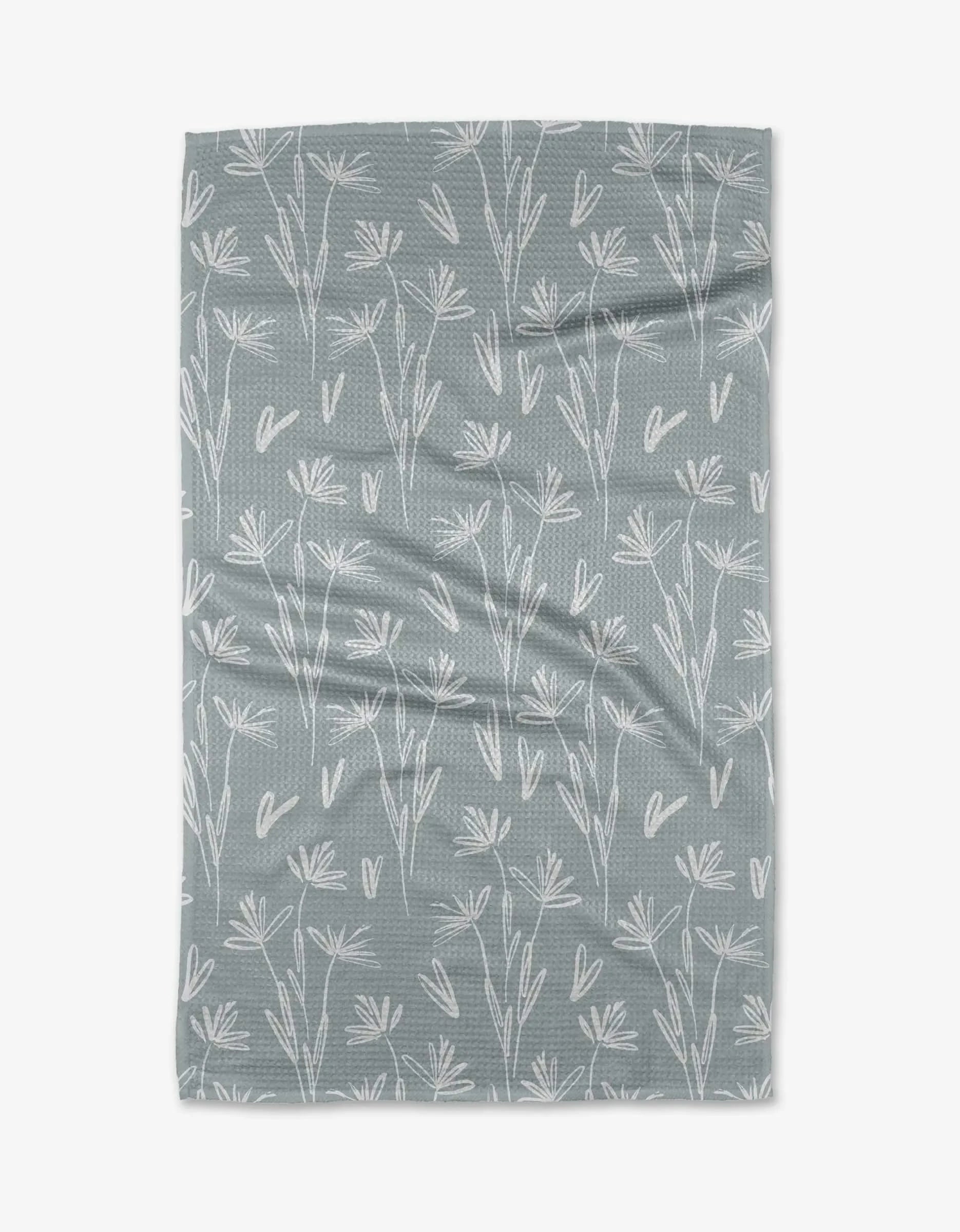 The Sketchbook Floral tea towel by Toboggan Avenue is a soft, greyish-blue kitchen towel with white hand-drawn floral patterns, made from recycled materials and shown flat with natural creases.