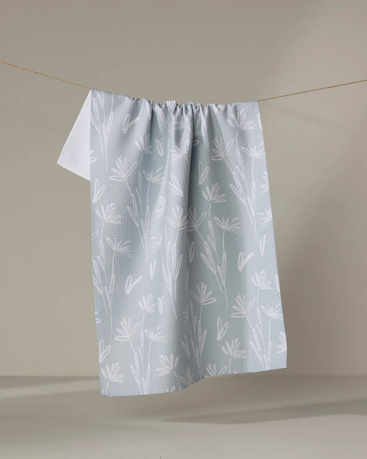 The Sketchbook Floral dish towel by Toboggan Avenue, featuring a light gray color with a white floral and leaf pattern, hangs on a string against a neutral background.