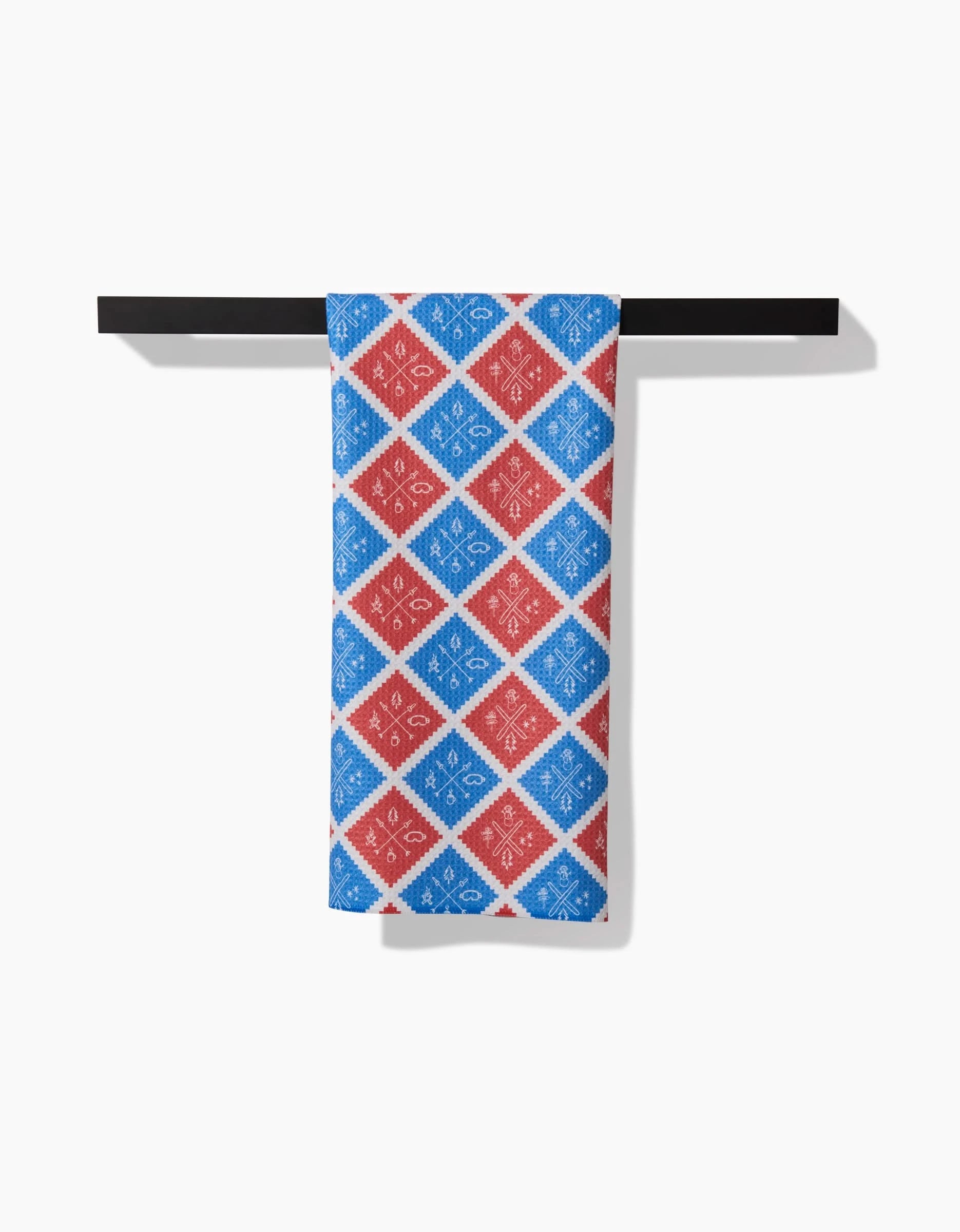 The Rosa Kichline Ski Patch tea towel, crafted from durable Geoweave fabric, features a red and blue diamond pattern with white snowflake-like designs and hangs neatly on a black bar against a plain white background.