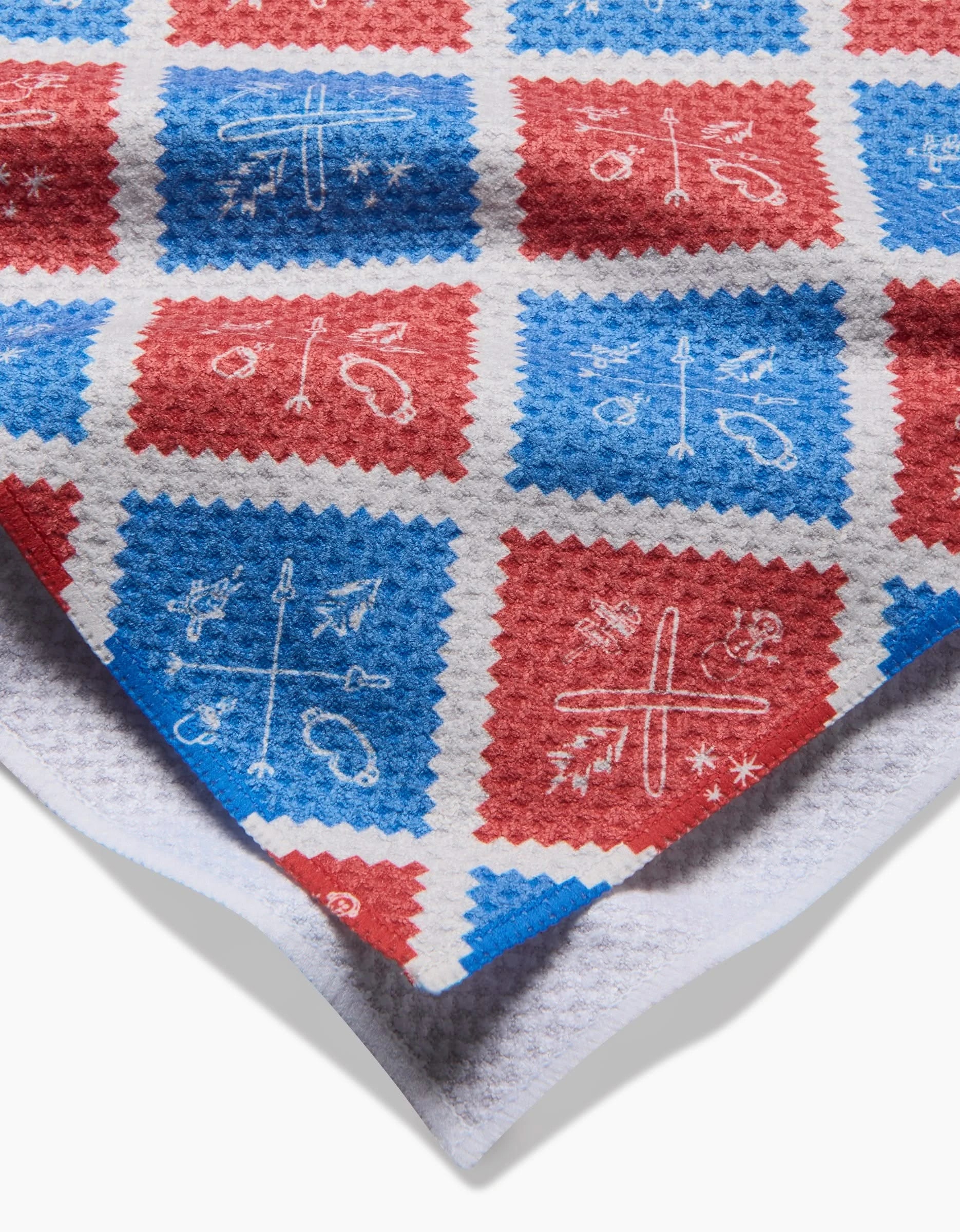 The Ski Patch by Rosa Kichline is a quick-drying tea towel made from recycled materials, featuring a red and blue checkered pattern with white ski-themed line drawings, and comes with one corner neatly folded over.