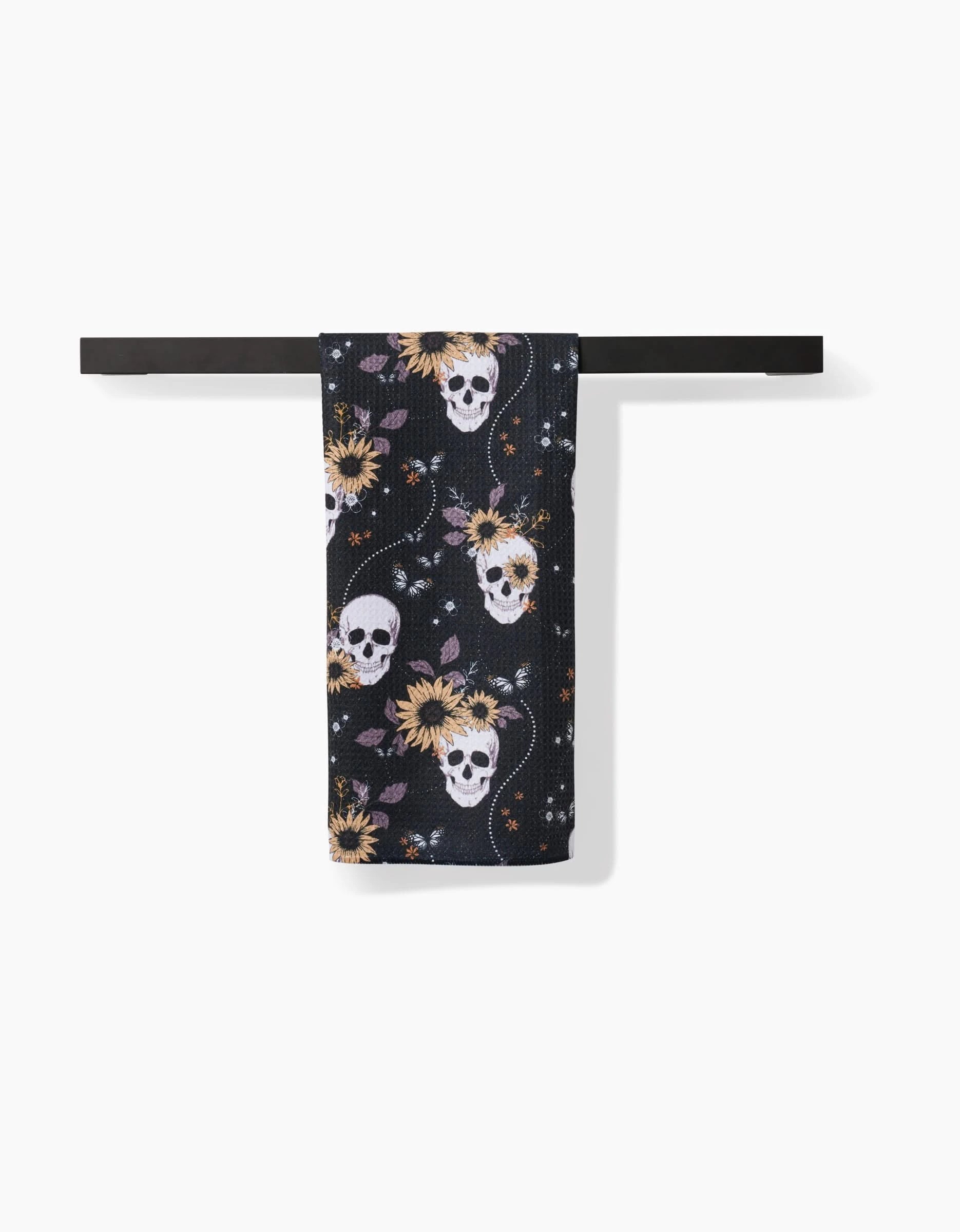 The Skulls In Bloom tea towel by Ashley Celena, made from recycled materials, showcases white skulls, yellow sunflowers, purple leaves, and orange stars on quick-drying black fabric—adding bold style to any space.