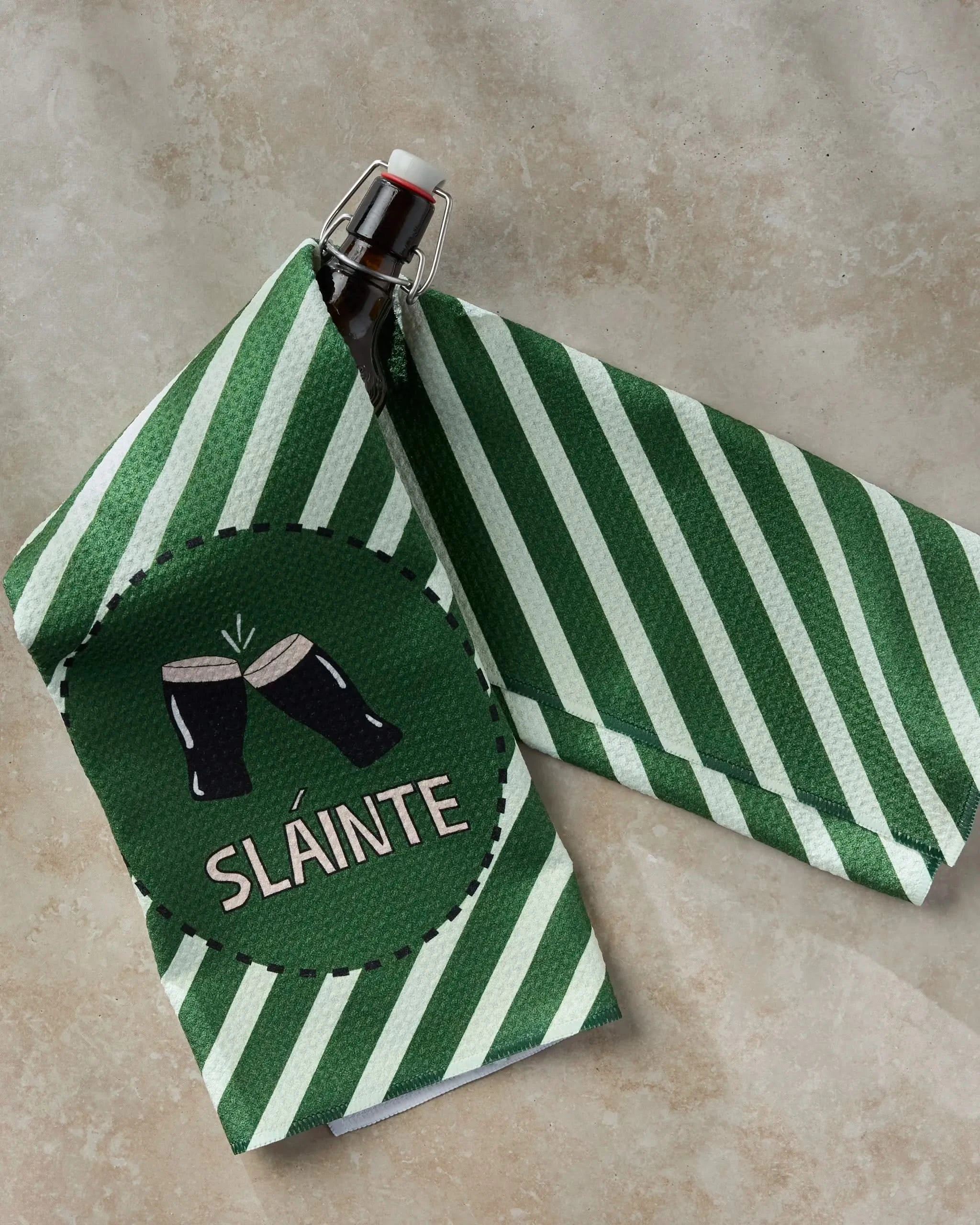 The Geometry "Slainte" tea towel, made from ultra-absorbent recycled materials, features green and white stripes, two clinking beer glasses, and the word SLÁINTE; it's shown draped over a swing-top bottle on a beige surface.