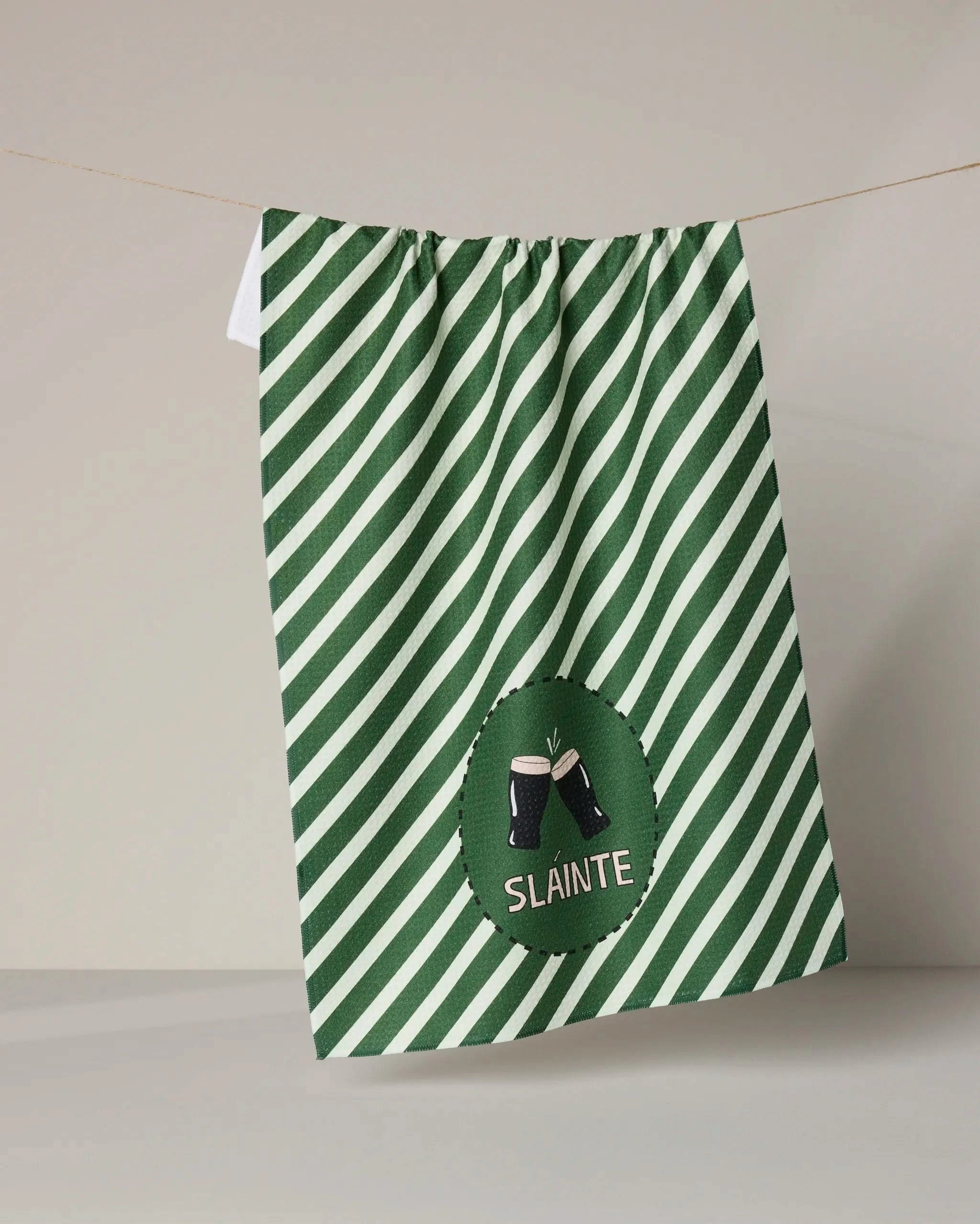 The Geometry "Slainte" tea towel features green and white diagonal stripes, is made from recycled materials, and displays an embroidered patch of two clinking beer glasses with "Sláinte" beneath them.