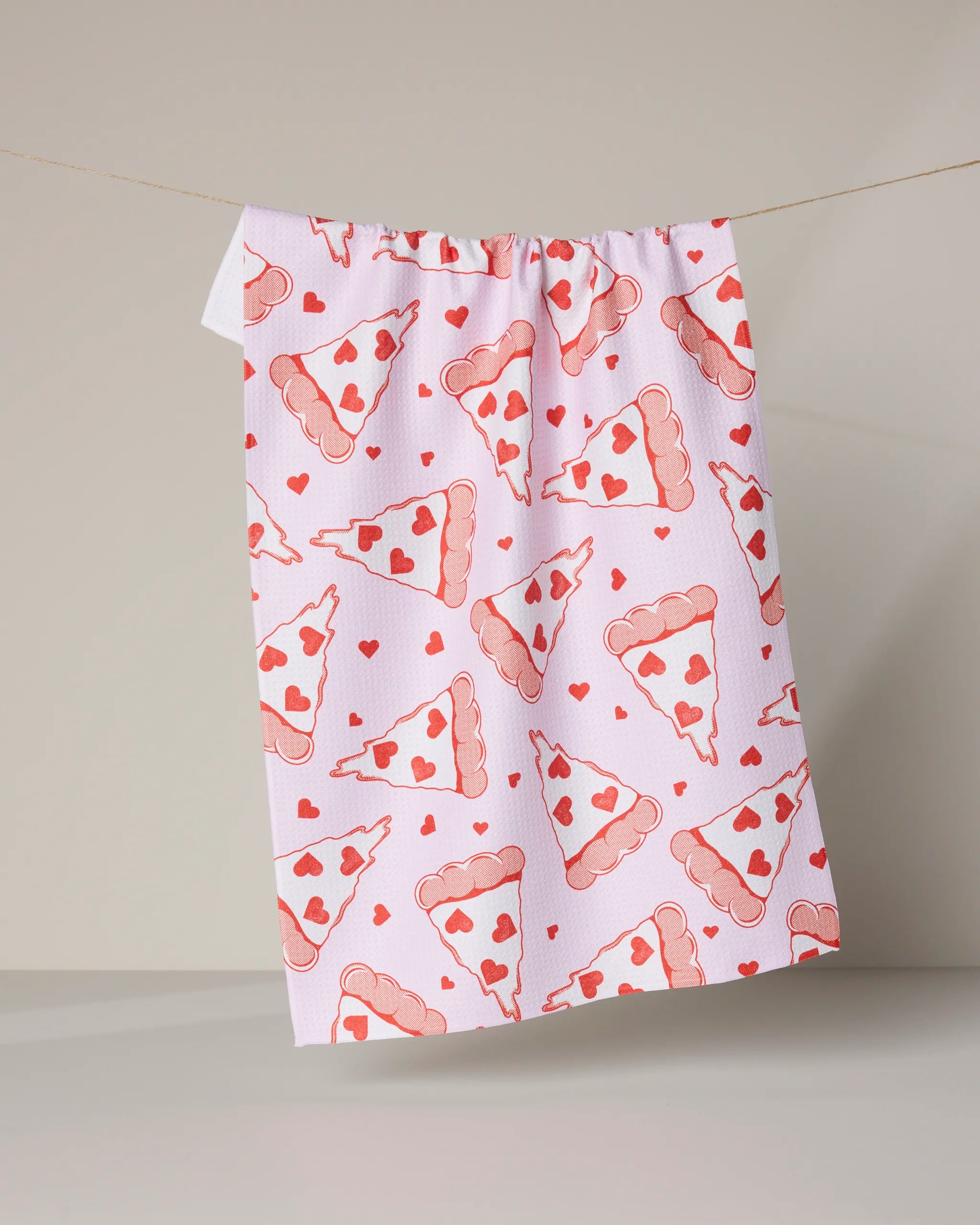 The Geometry "Slice of Heaven" tea towel, featuring pink pizza slices and red hearts, hangs on a clothesline against a neutral background.