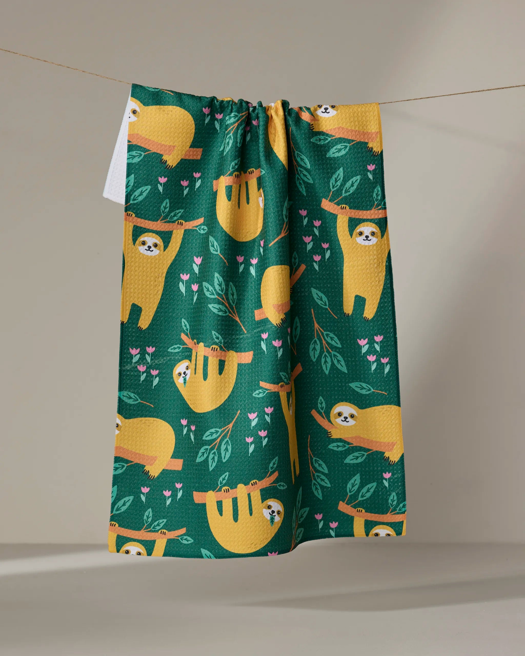 The "Sloths" tea towel by Genna Blackburn is green, made from recycled materials, and features playful yellow sloths with pink flowers and leaves, hanging from branches. It's shown on a clothesline against a neutral background.