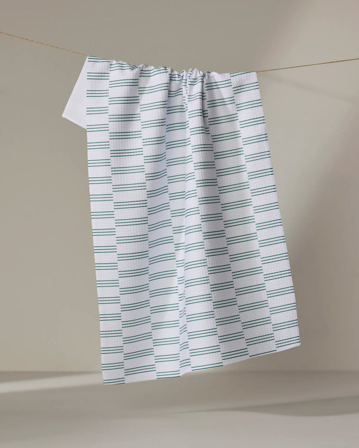 A Small Stripe - Evergreen dish towel by The Buy Guide, featuring blue and gray horizontal stripes, hangs on a clothesline against a beige background.