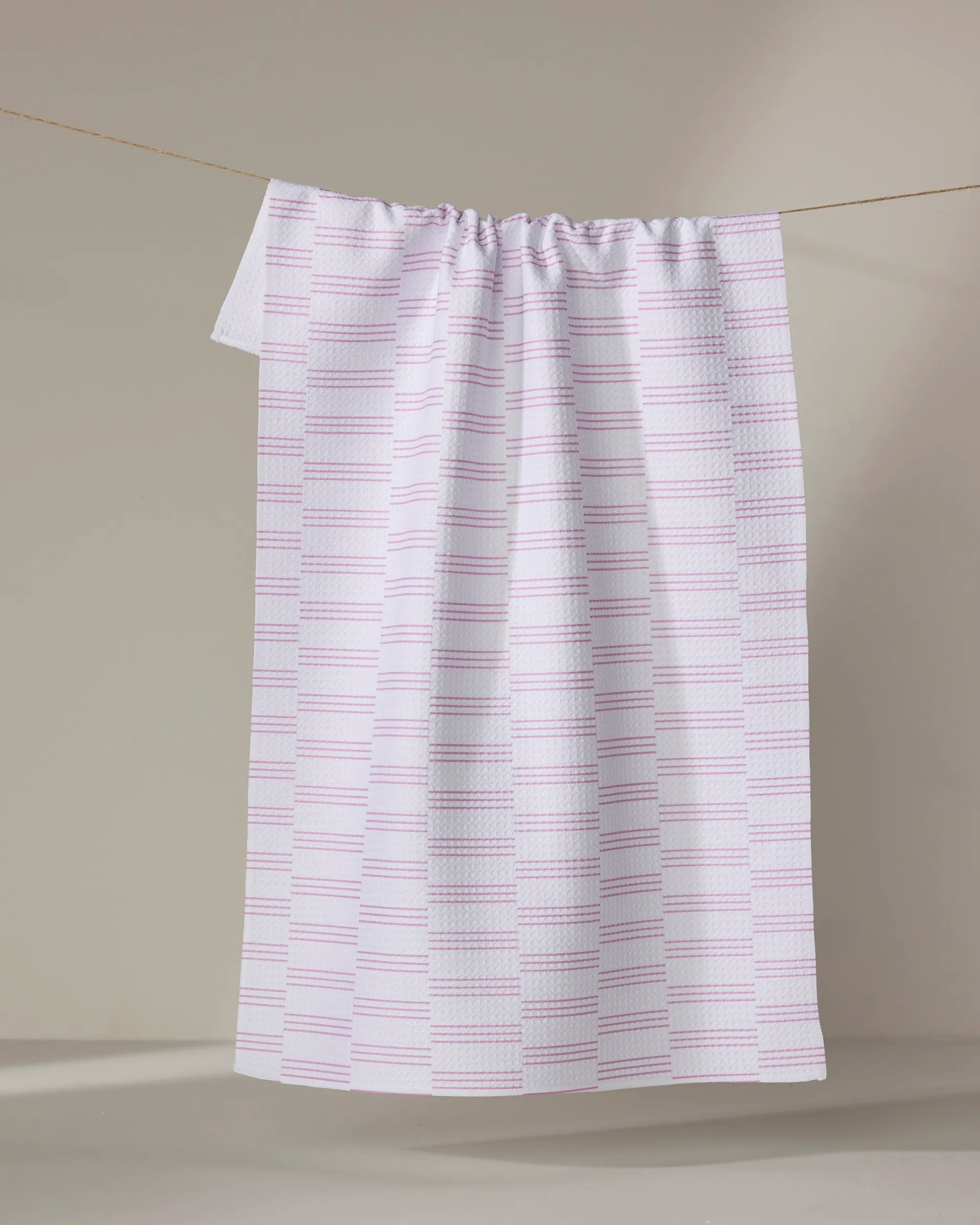 The Buy Guide's 'Small Stripe - Pink' ultra-absorbent white towel with a pink and purple grid pattern hangs neatly on a clothesline against a neutral beige background.