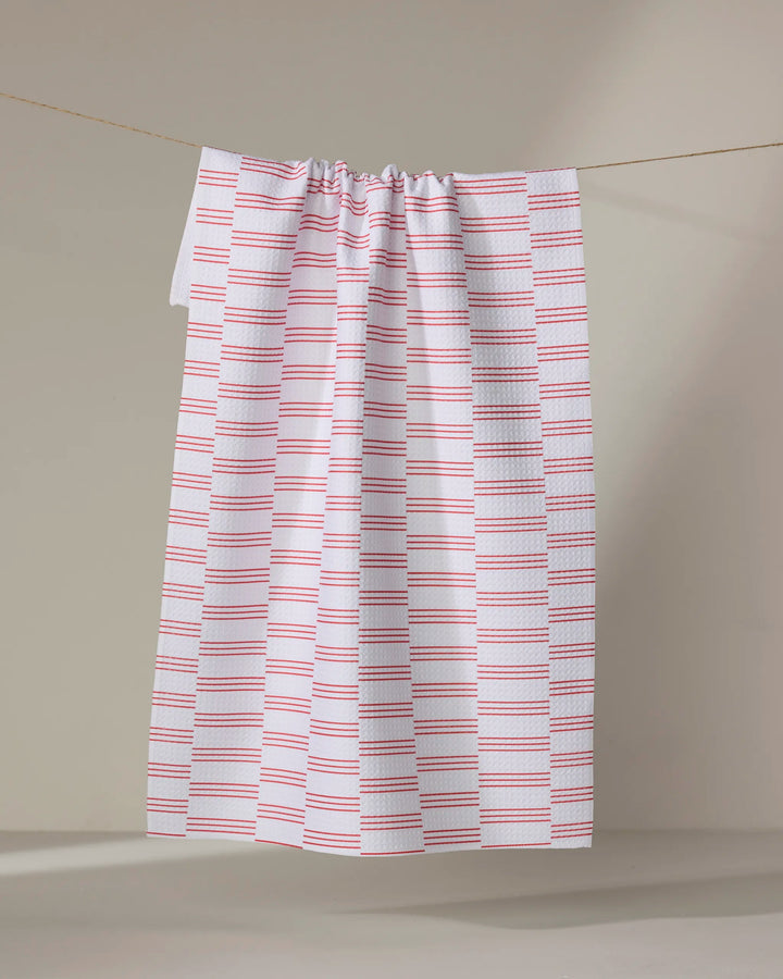 The Buy Guide's Small Stripe - Red tea towel, featuring evenly spaced horizontal red stripes on white, hangs from a string against a plain, neutral background.