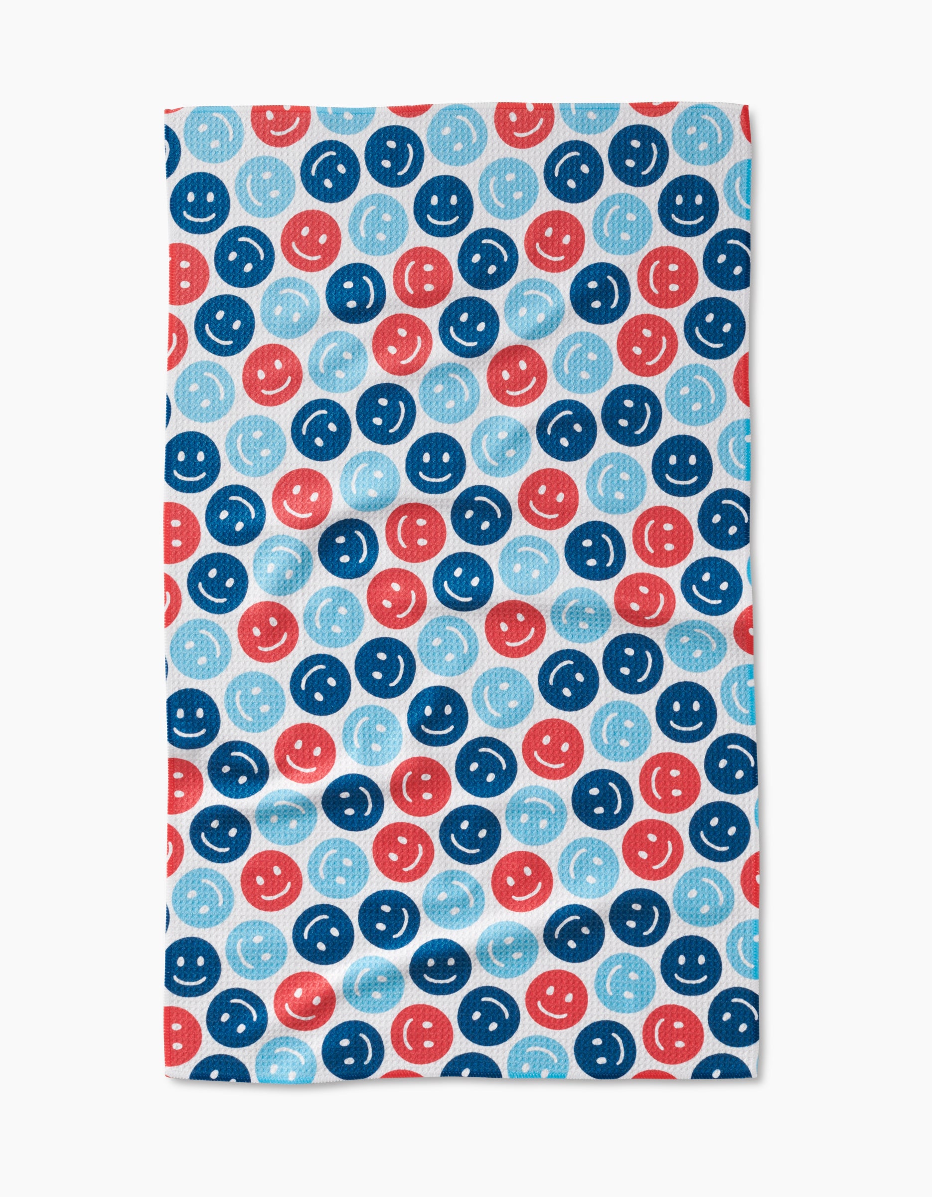 The Smiley Face July tea towel by Hufton Studio is made from recycled materials and features a pattern of blue, red, and light blue smiley faces with simple round eyes and curved mouths on a white background.