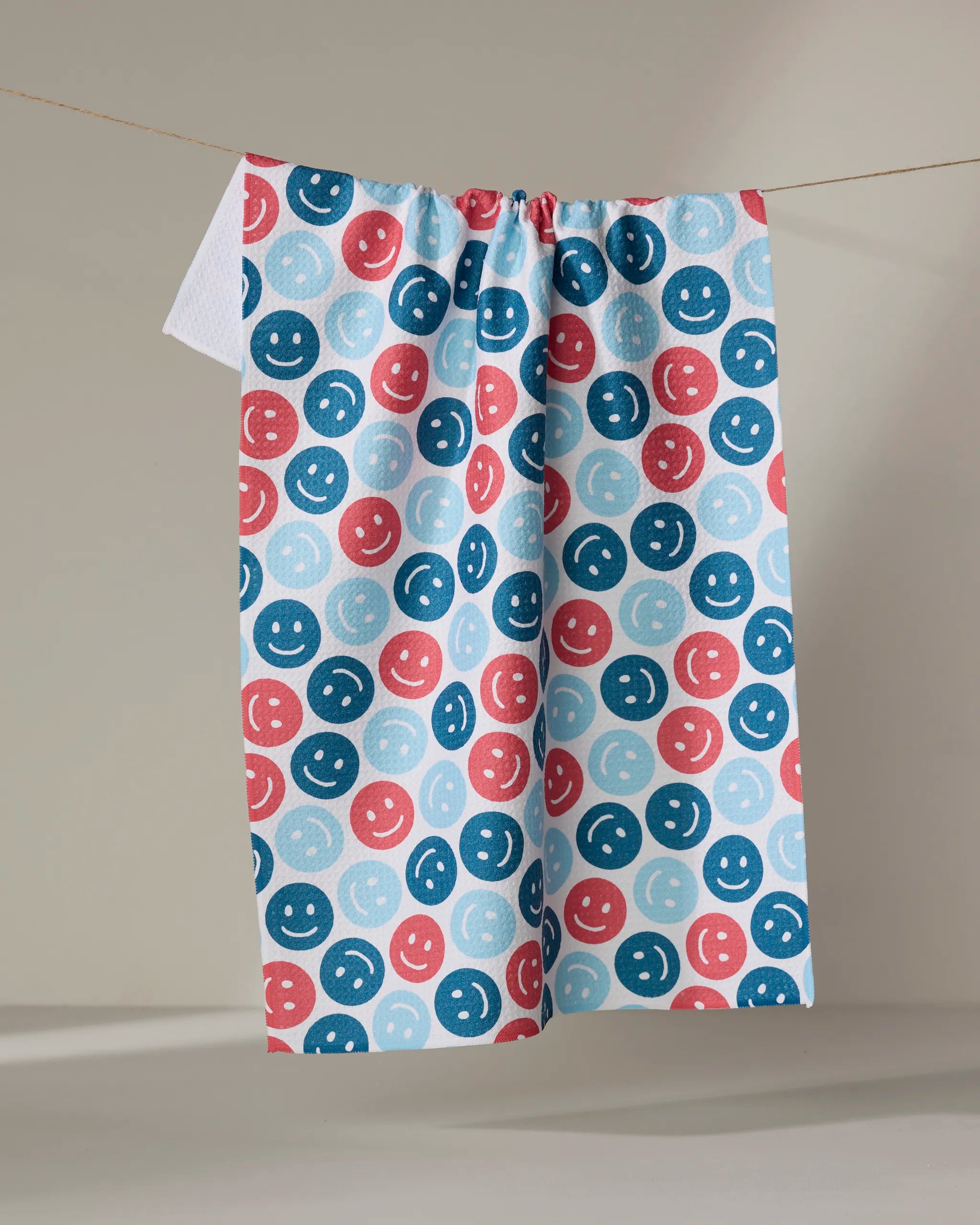 The Hufton Studio "Smiley Face July" tea towel, featuring red, blue, and light blue smiley faces, hangs on a clothesline against a neutral background.