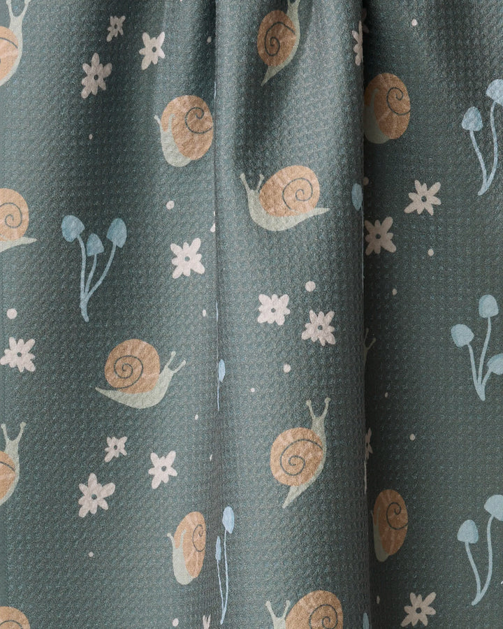A close-up of the Snail Gathering dish towel by Amberleigh Adoff shows its absorbent green fabric with a playful print of snails, white flowers, and blue mushrooms.