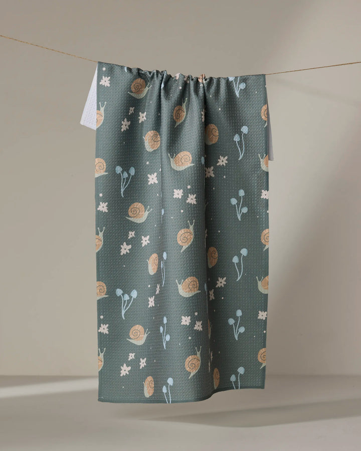 The Snail Gathering kitchen towel by Amberleigh Adoff, featuring beige snails, light blue flowers, and white geometric shapes on dark teal, hangs from a taut string against a neutral wall.