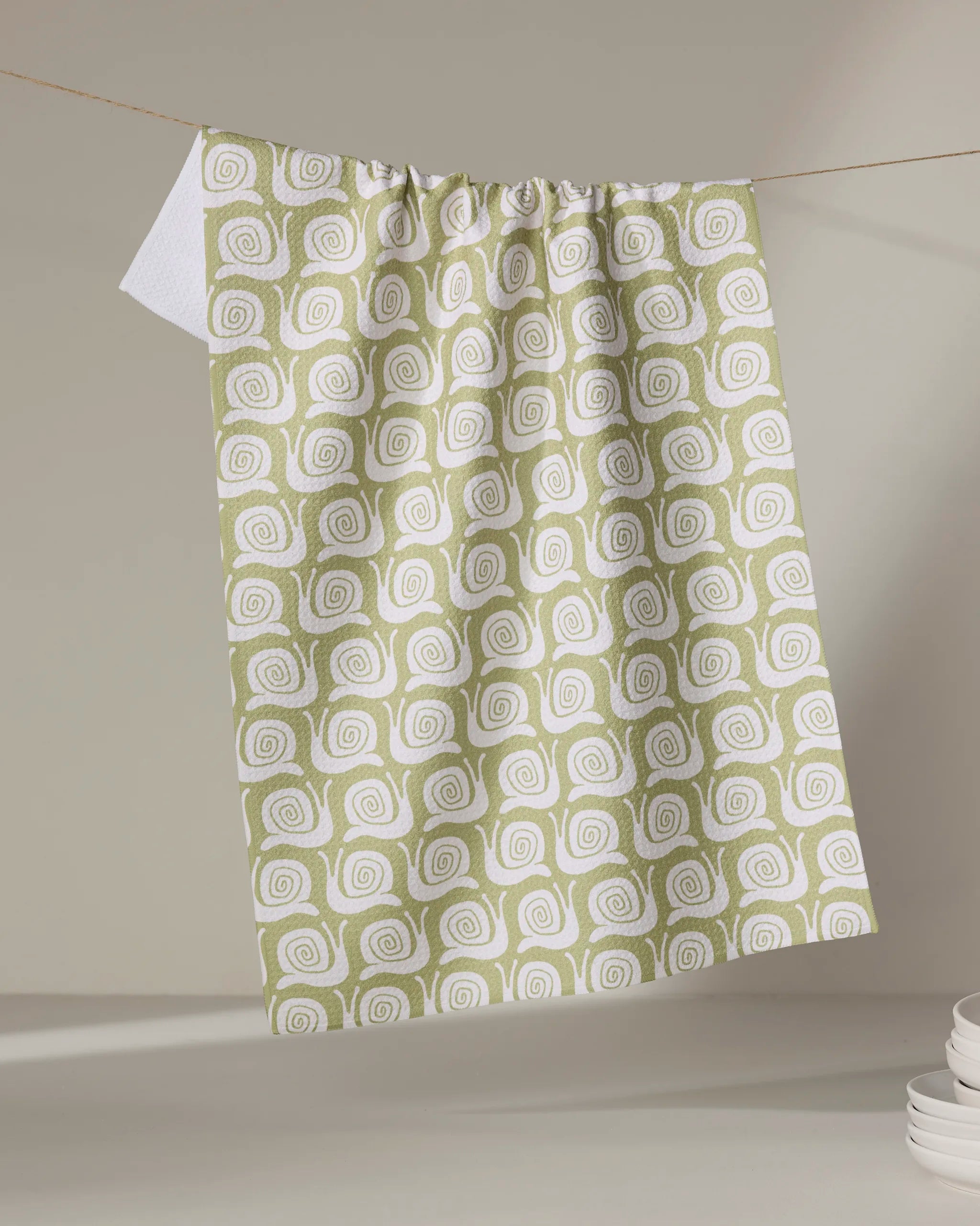 The Kayla Ann Snail Party kitchen towel, featuring a green and white spiral design made from recycled materials, hangs on a clothesline with white dishes partially visible against a neutral background.