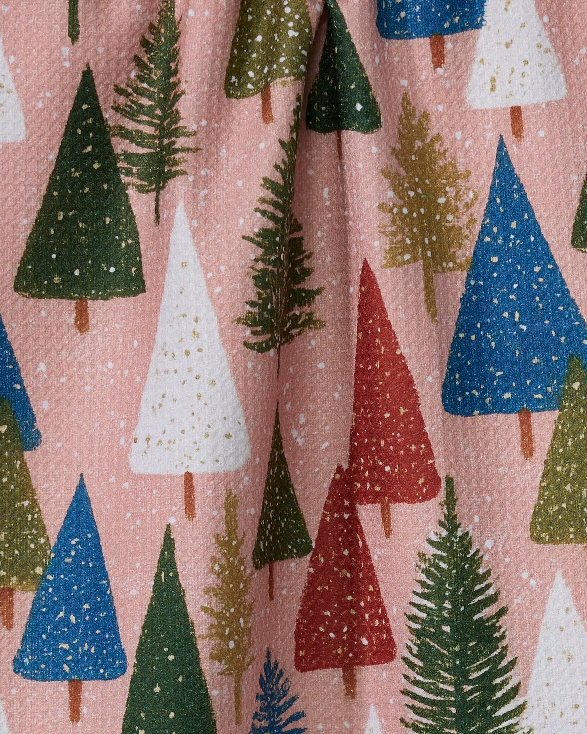 The Geometry "Snowdrift Pines" tea towel features green, red, blue, and white Christmas trees with brown trunks on a light pink textured background, accented by small white dots like falling snow.