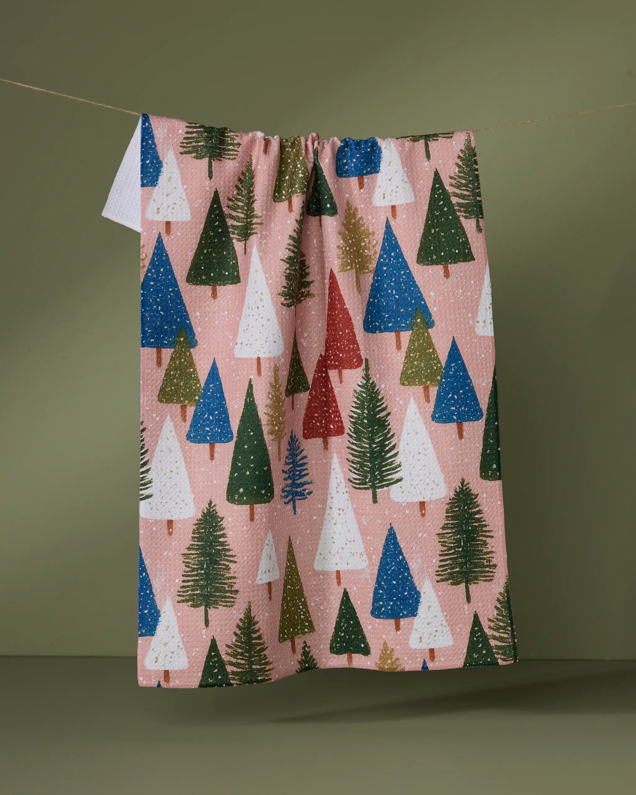 The Geometry "Snowdrift Pines" dish towel features absorbent fabric adorned with green, white, red, and blue Christmas trees on a pink background, shown hanging on a line against an olive-green backdrop.
