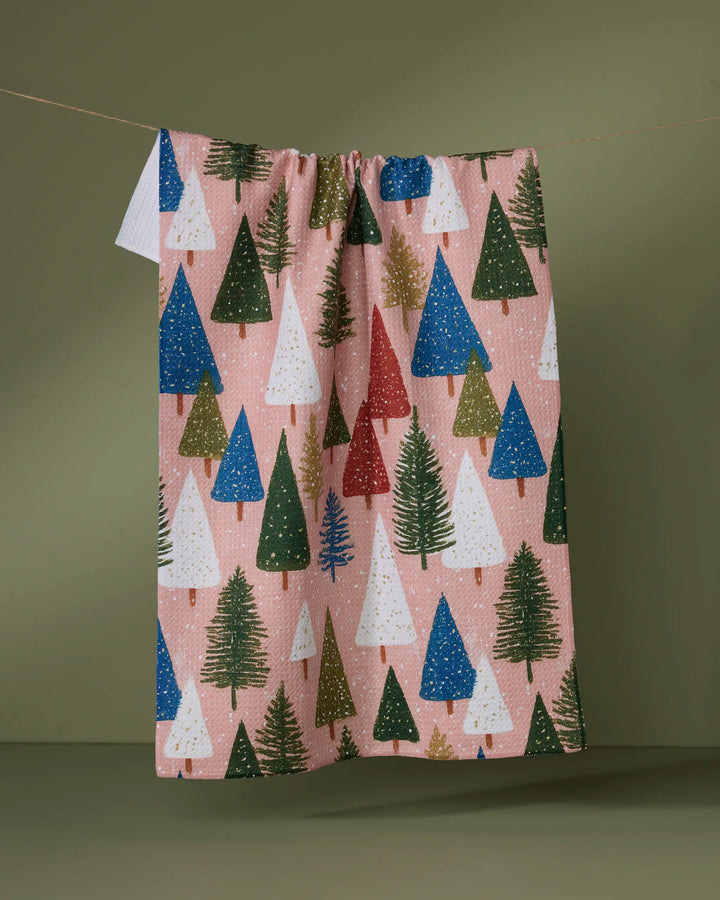 The Geometry "Snowdrift Pines" dish towel features absorbent fabric adorned with green, white, red, and blue Christmas trees on a pink background, shown hanging on a line against an olive-green backdrop.