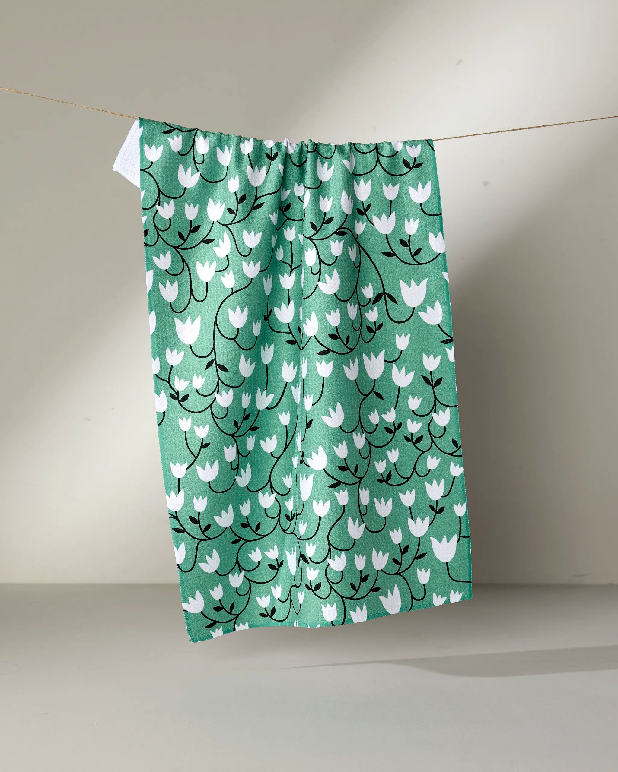 The Snowdrop tea towel by Rachel Parker features a white and black floral pattern on green, made from recycled materials, and is displayed hanging on a thin string against a neutral background.
