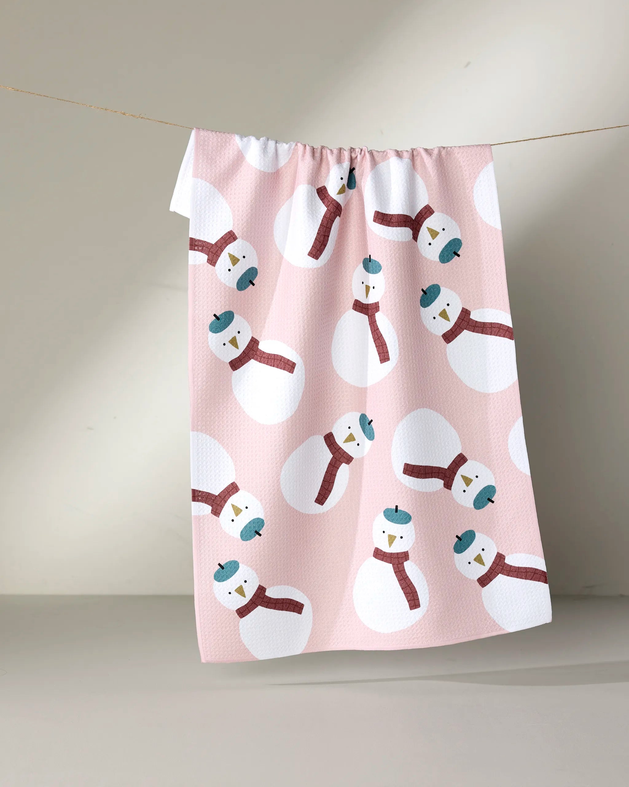 The "Snowman" tea towel by Jennifer Bouron features cute snowmen with blue hats and red scarves on pink, made from ultra-absorbent recycled materials, and hangs on a clothesline against a neutral background.
