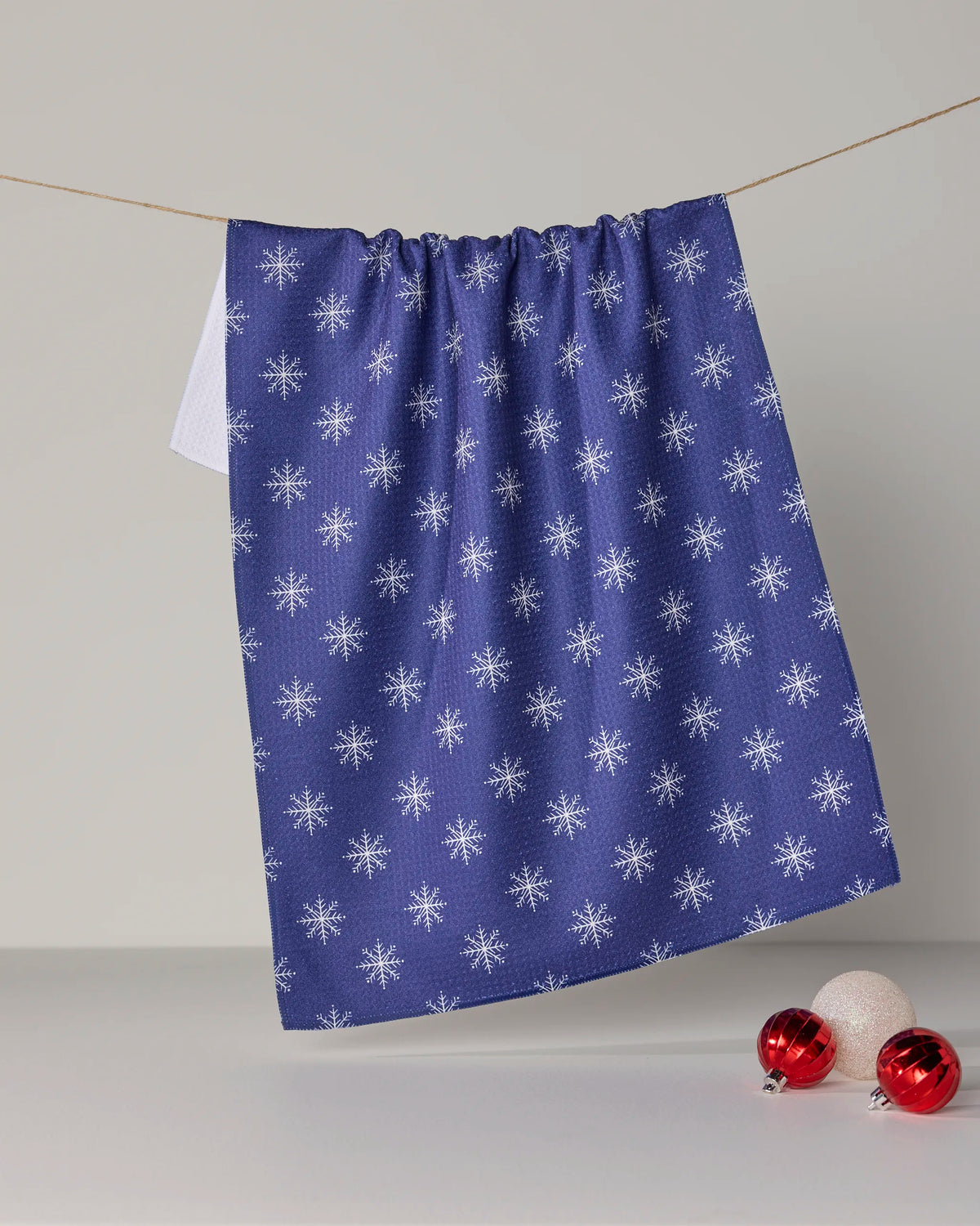 A Geometry “Snowy” tea towel, crafted from recycled materials and featuring a blue design with white snowflake patterns, hangs on a string. Two red ornaments and one white ornament rest on the white surface below.