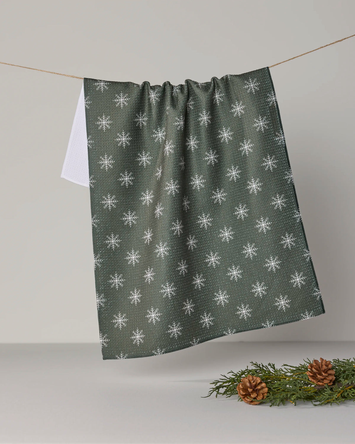 The “Snowy” tea towel by Geometry, featuring green ultra-absorbent fabric adorned with white snowflake patterns, hangs on a string. A white towel is partially visible behind it, while pine cones and evergreen sprigs in the bottom right corner accentuate its eco-friendly construction from recycled materials.