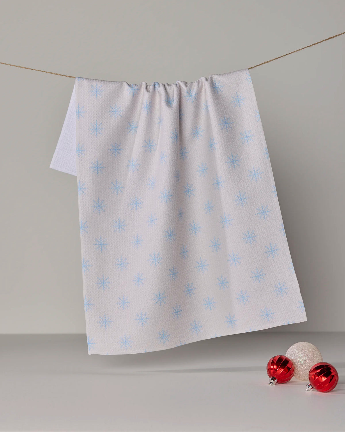 The Geometry Snowy tea towel, featuring light blue snowflake patterns on a white background and crafted from recycled materials, hangs on a string above two red ornaments and one white ornament displayed on the surface below.