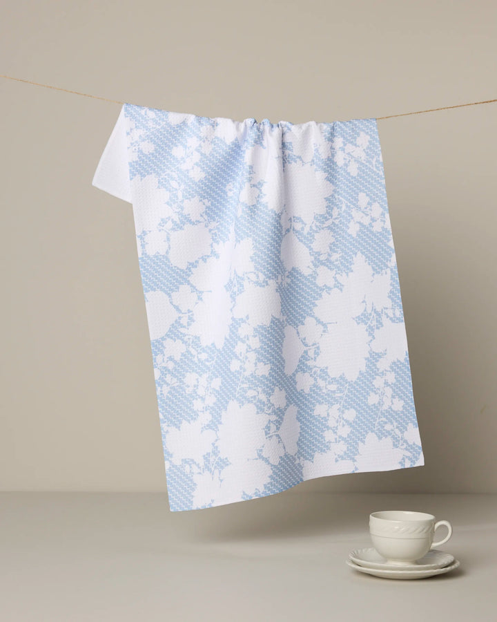 The Geometry "Soft Bloom" tea towel, featuring a light blue and white pattern and made from recycled materials, hangs on a string with a white teacup and saucer set nearby against a plain background.