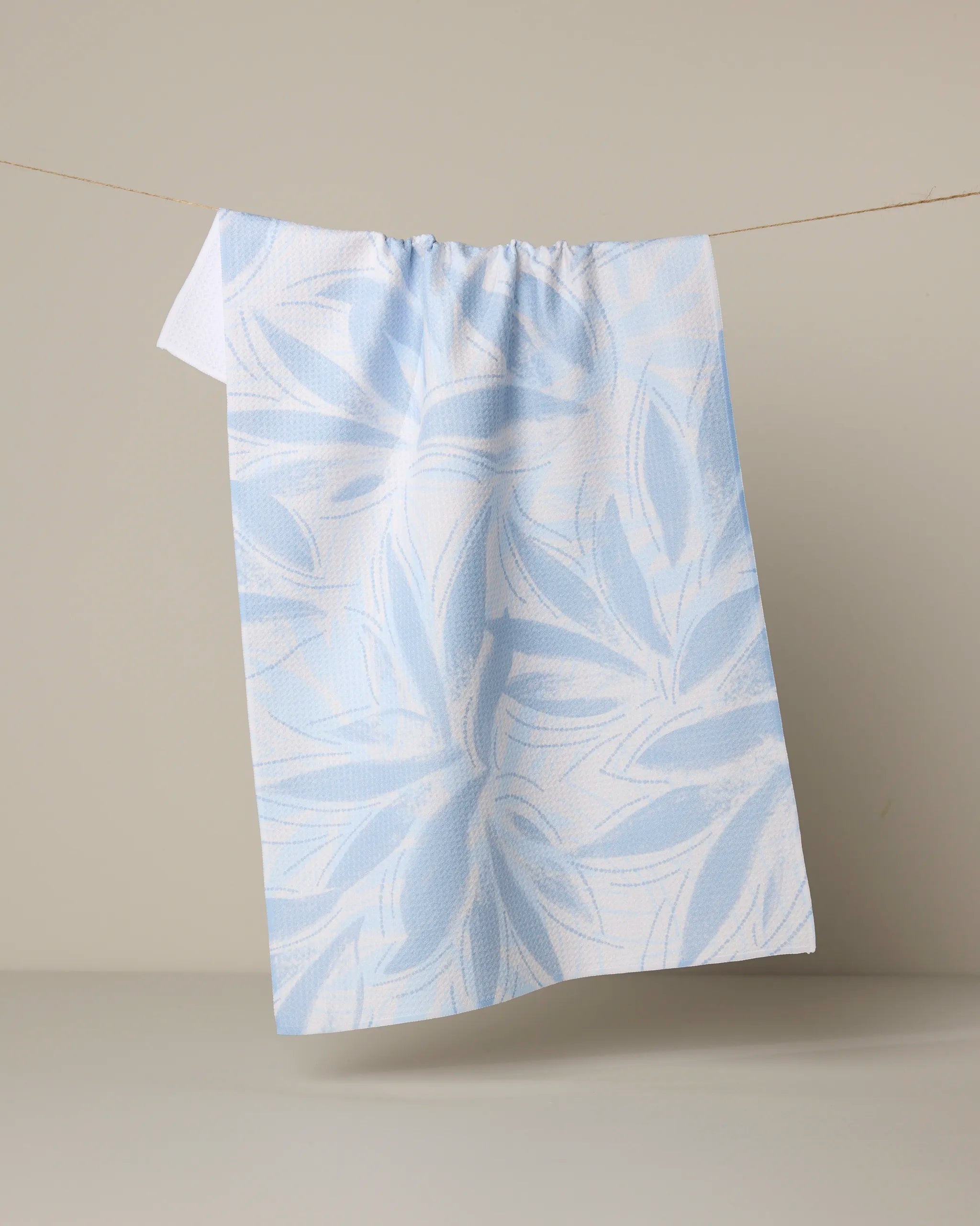 The Geometry "Soft Breeze" tea towel, featuring a white and light blue leaf pattern, hangs on a thin string against a plain beige background.