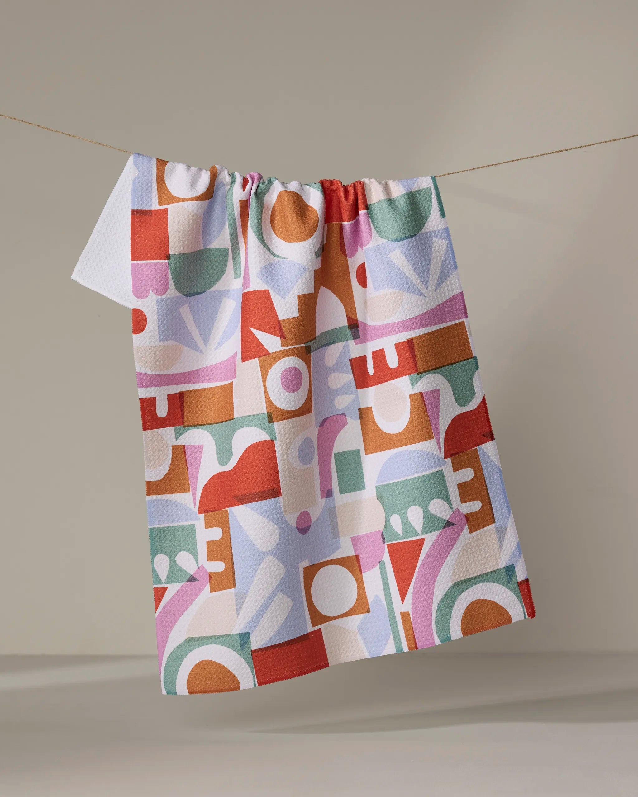 The "Soft Cutouts Garden" kitchen towel by Claire Iglesias, featuring an abstract geometric pattern in red, green, orange, and purple, hangs on a clothesline against a plain beige background.