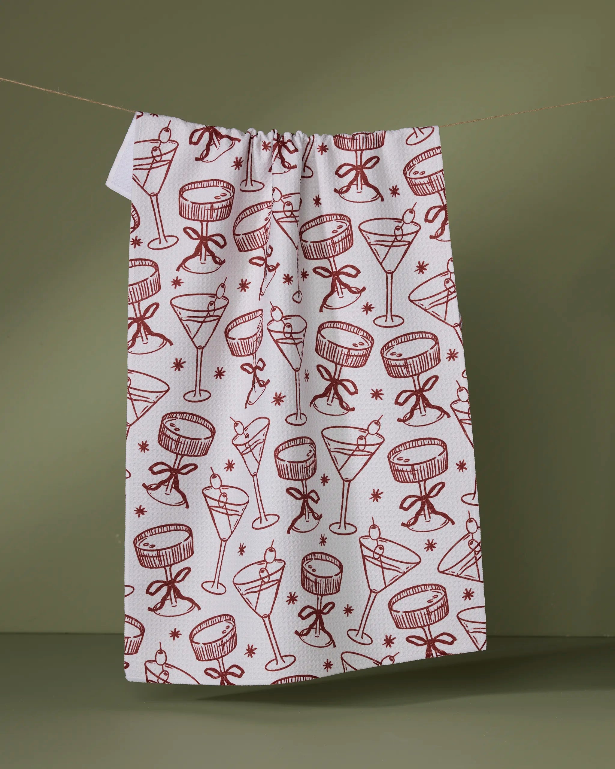 The Geometry Soiree tea towel, with quick-drying fabric and red cocktail glass, star, and bow illustrations, hangs from a line against an olive green background.
