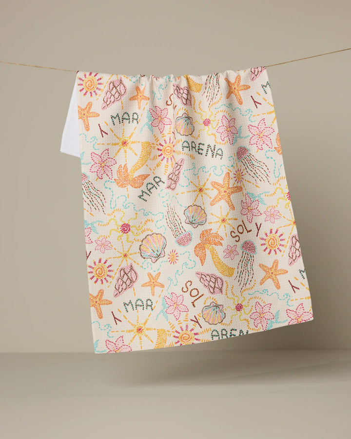 The Sol y Mar kitchen towel by Geometry hangs on a line, displaying playful starfish, seashells, and words like MAR, ARENA, and SOL in pastel hues—a beach-themed design made from recycled materials.