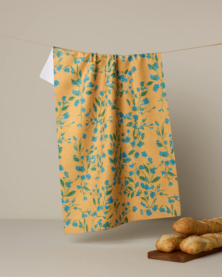 The Geometry Soleado tea towel, featuring a yellow ultra-absorbent fabric with a blue floral pattern, hangs against a beige background as a wooden board of baguettes sits in the lower right corner.