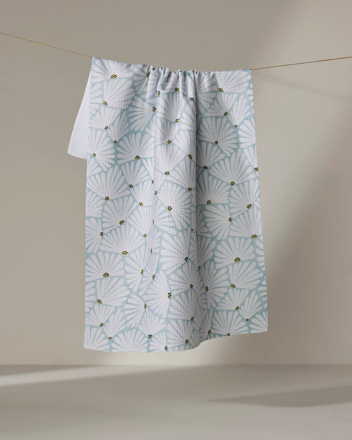 The Solie Teal tea towel by Amy MacCready, crafted from recycled materials, features a quick-drying white fabric with a light blue scallop shell pattern and small yellow bee accents, hanging against a neutral background.