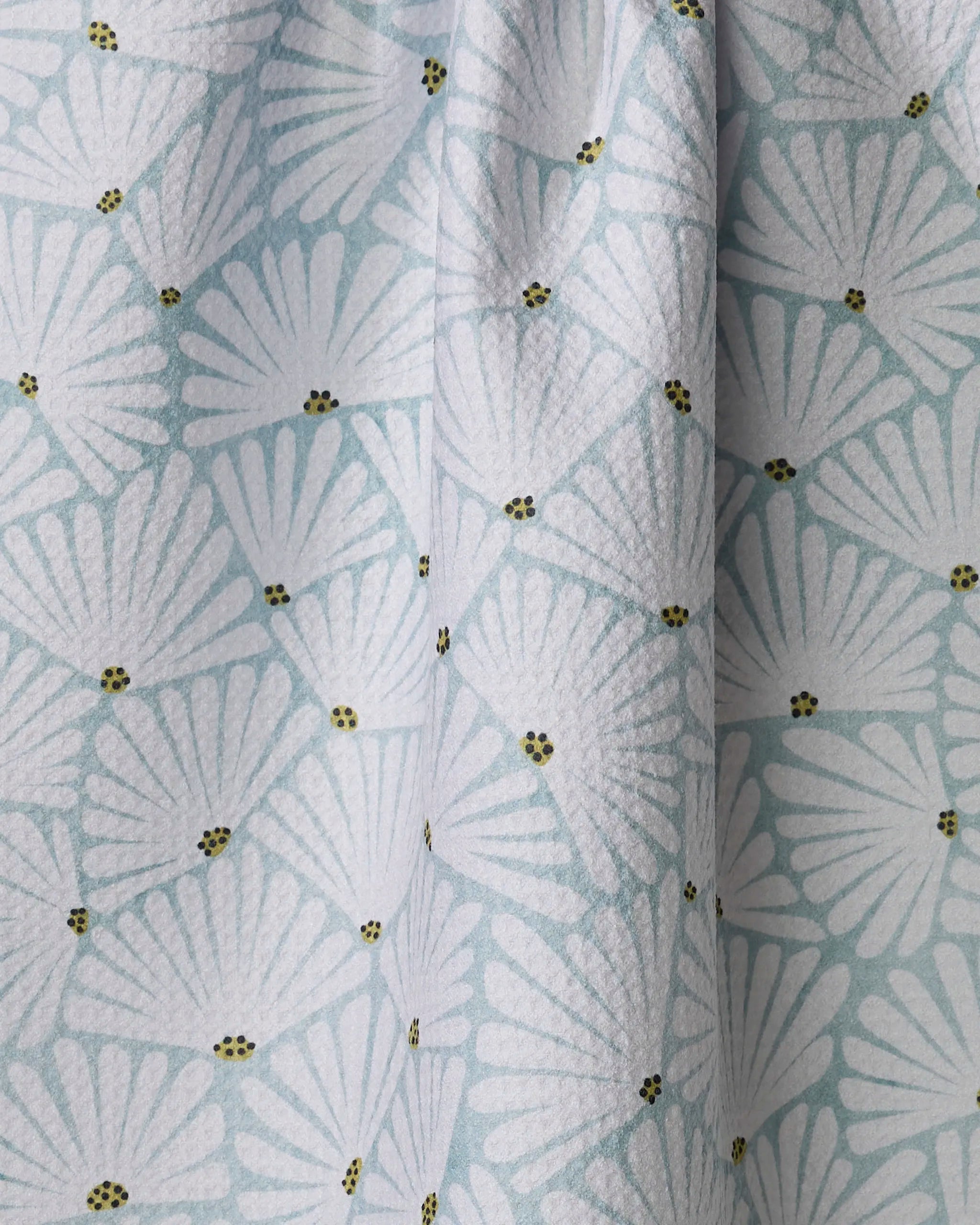 The Solie Teal tea towel by Amy MacCready is made from recycled materials and features a light blue color with white fan patterns plus yellow and black dots. Its fabric drapes softly, forms gentle folds, and dries quickly for daily use.